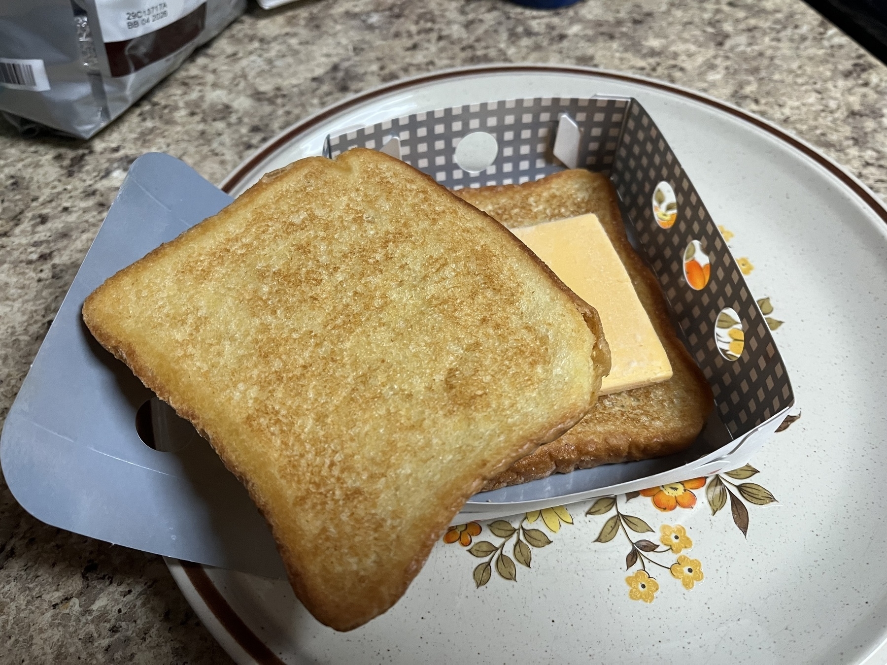 Auto-generated description: A piece of toast with cheese sits inside a decorative, partially open sandwich box on a floral-patterned plate.