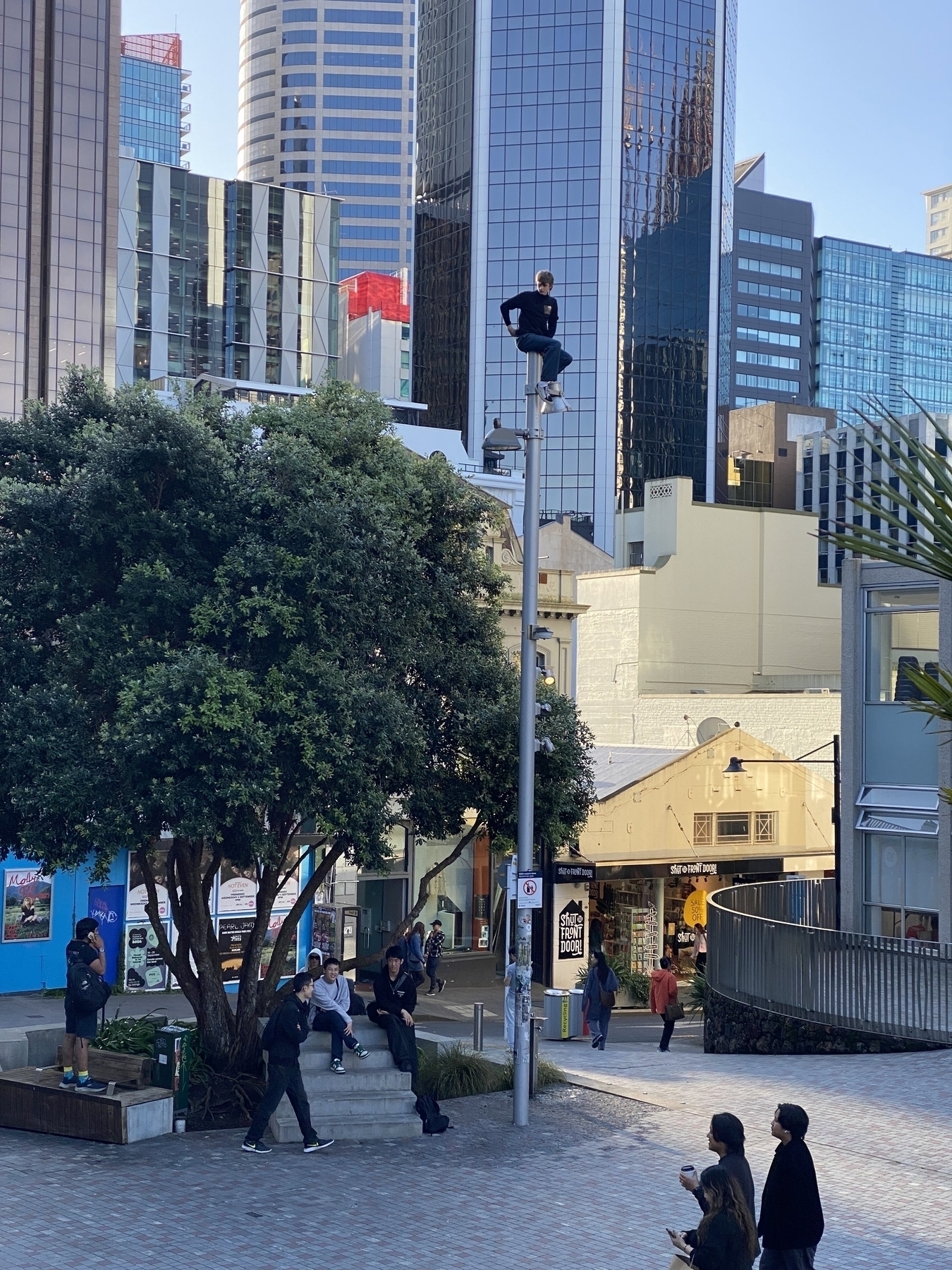 A person is standing on top of a pole overlooking a plaza in Auckland with CBD Skyscrapers in the background.