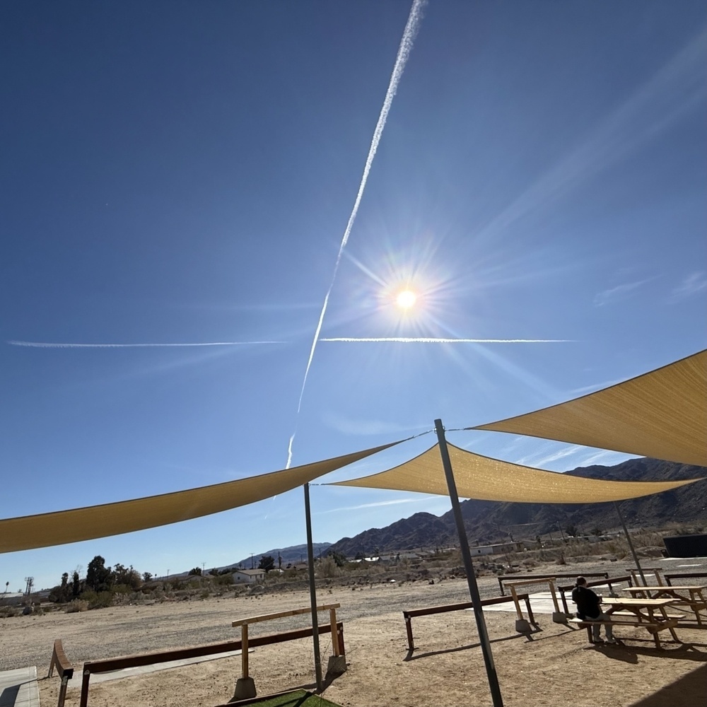 A sunny outdoor area features triangular shade sails, a crisscrossing jet trail in the sky, and a mountainous backdrop.