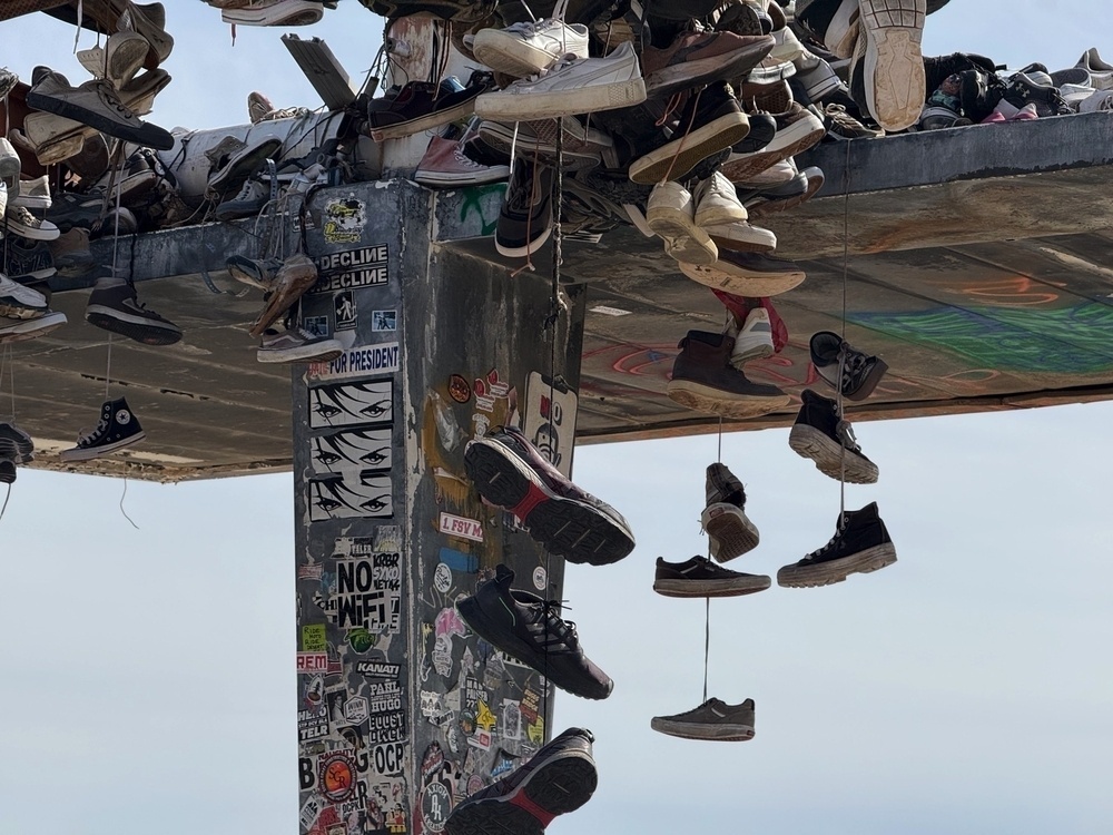 Numerous sneakers are hanging by their laces from a structure covered in stickers and graffiti.