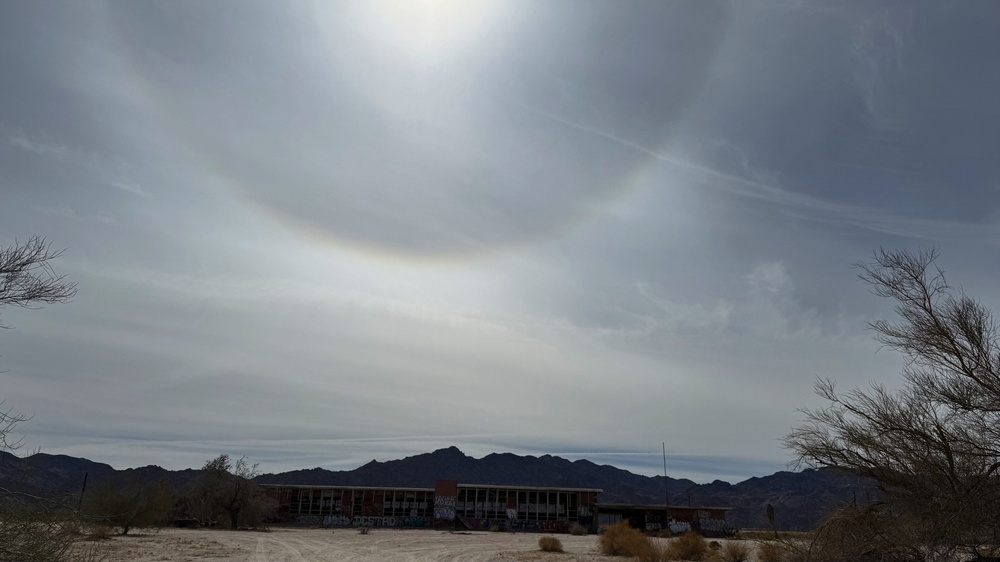 A large, bright halo is visible in the cloudy sky above a deserted landscape with a building surrounded by barren trees and distant mountains.