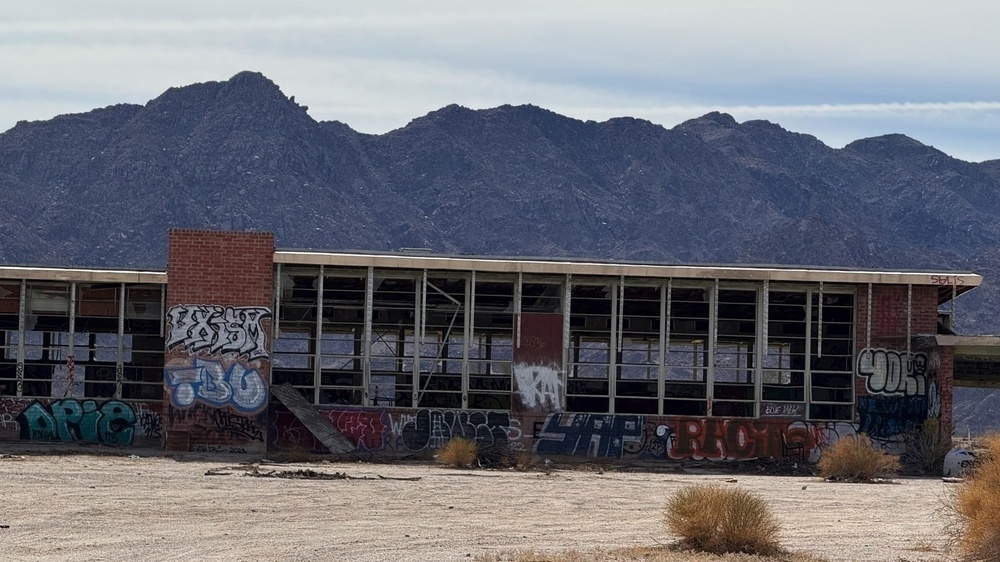 An abandoned building covered in graffiti stands in a barren area with mountains in the background.