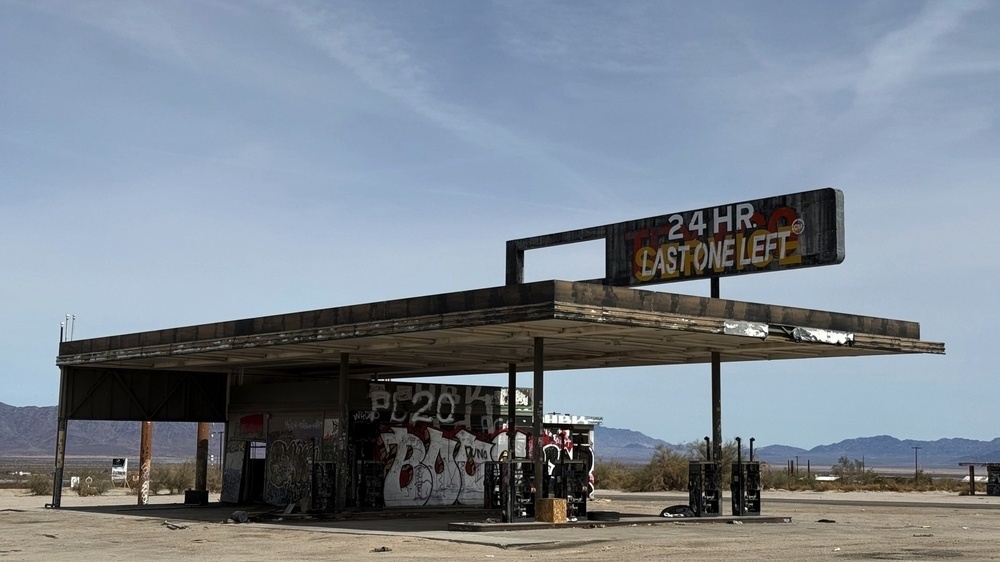 A deserted and dilapidated gas station stands under an empty sky with graffiti on its walls and a partially broken sign.