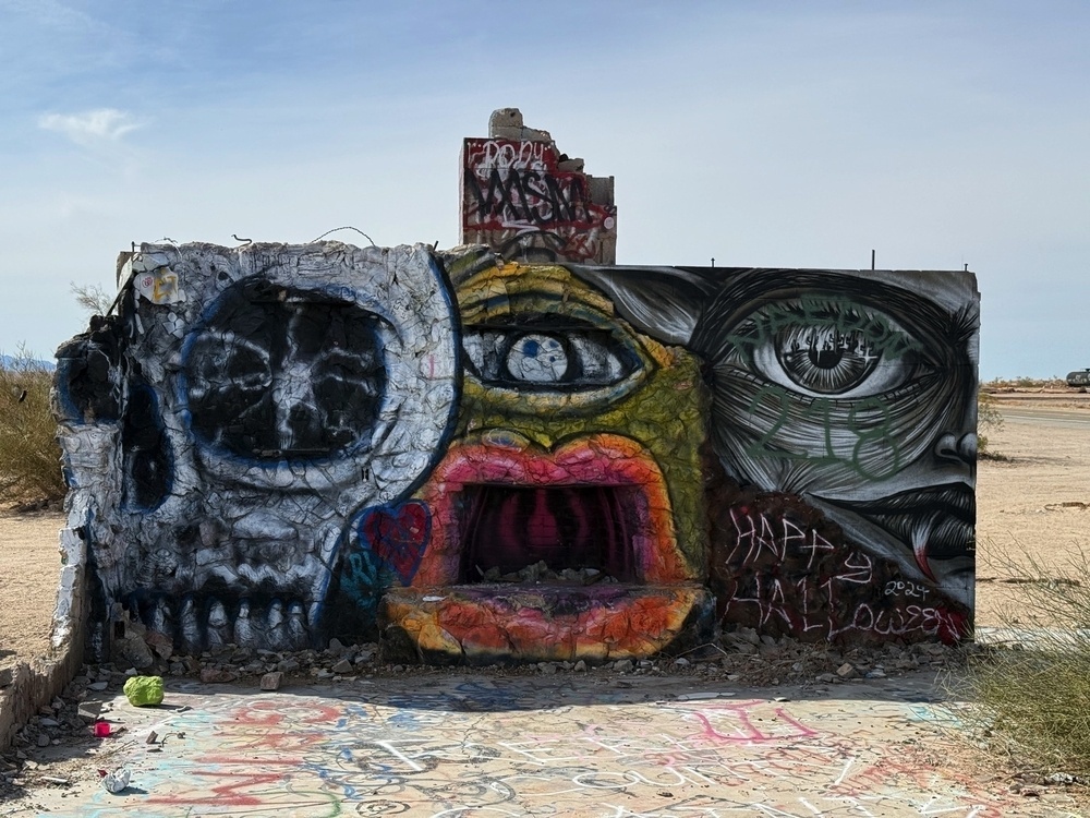 A graffiti-covered wall in a desert area features a skull, a colorful abstract face, and detailed eyes.