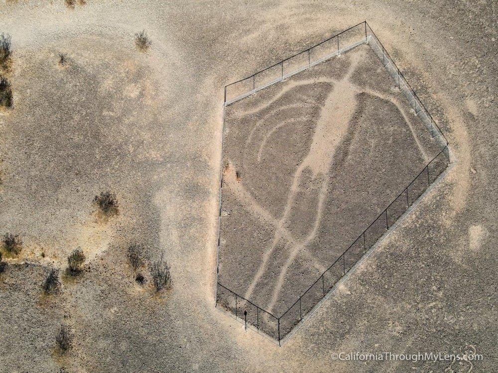 An aerial view shows a fenced, large geoglyph of a human figure in a barren landscape.