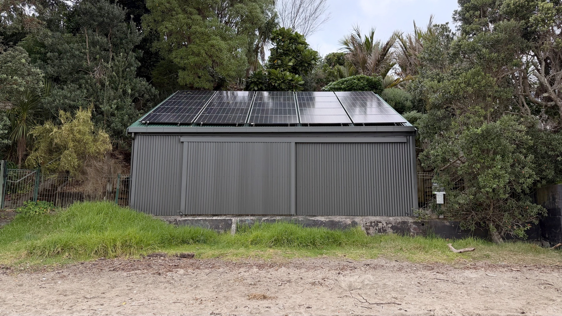 A small structure with a dark roof covered in solar panels sits nestled among a dense backdrop of lush green trees.