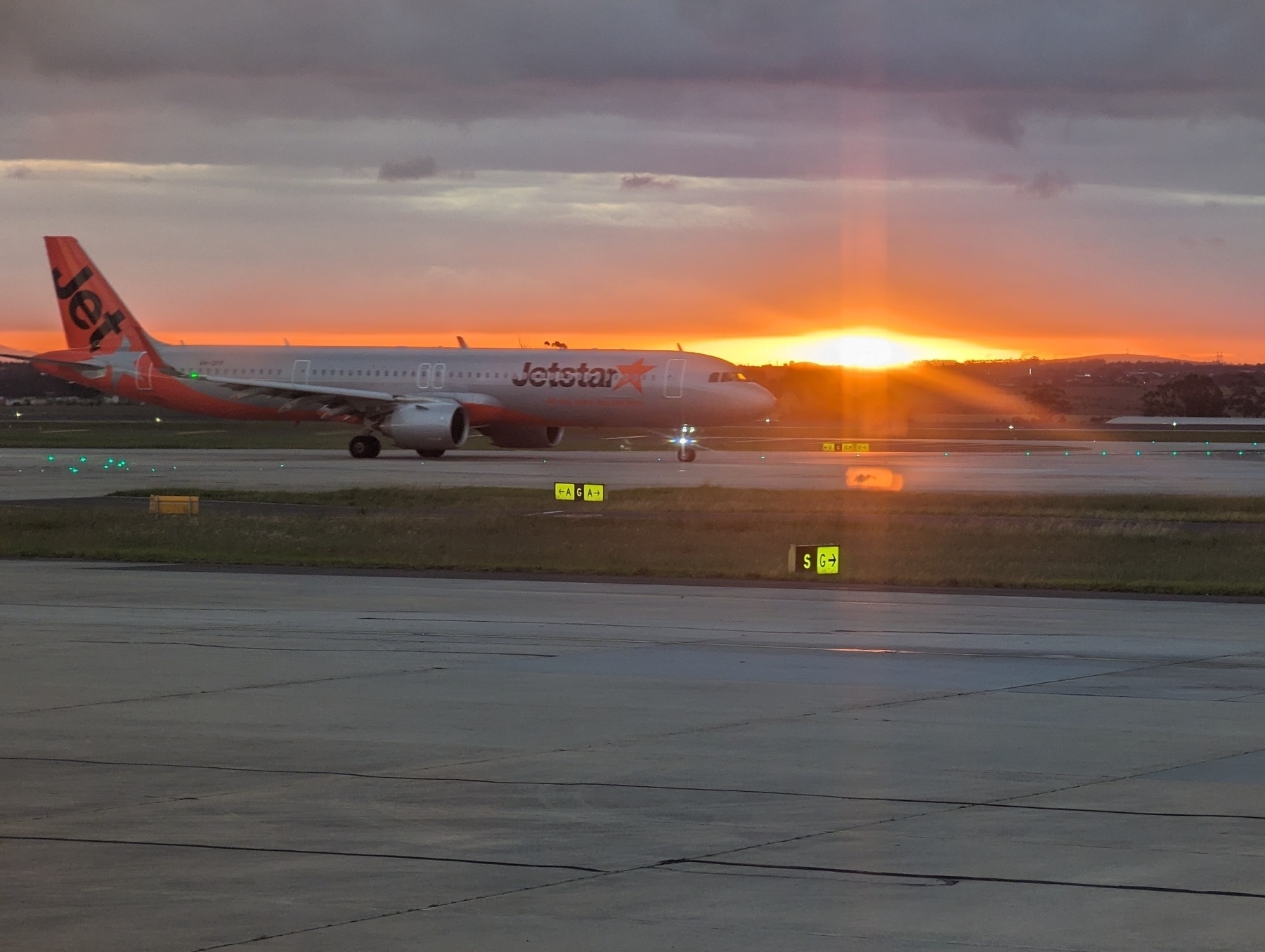 Airplane taxing at an airport with an orange sunset flaring near the nose.