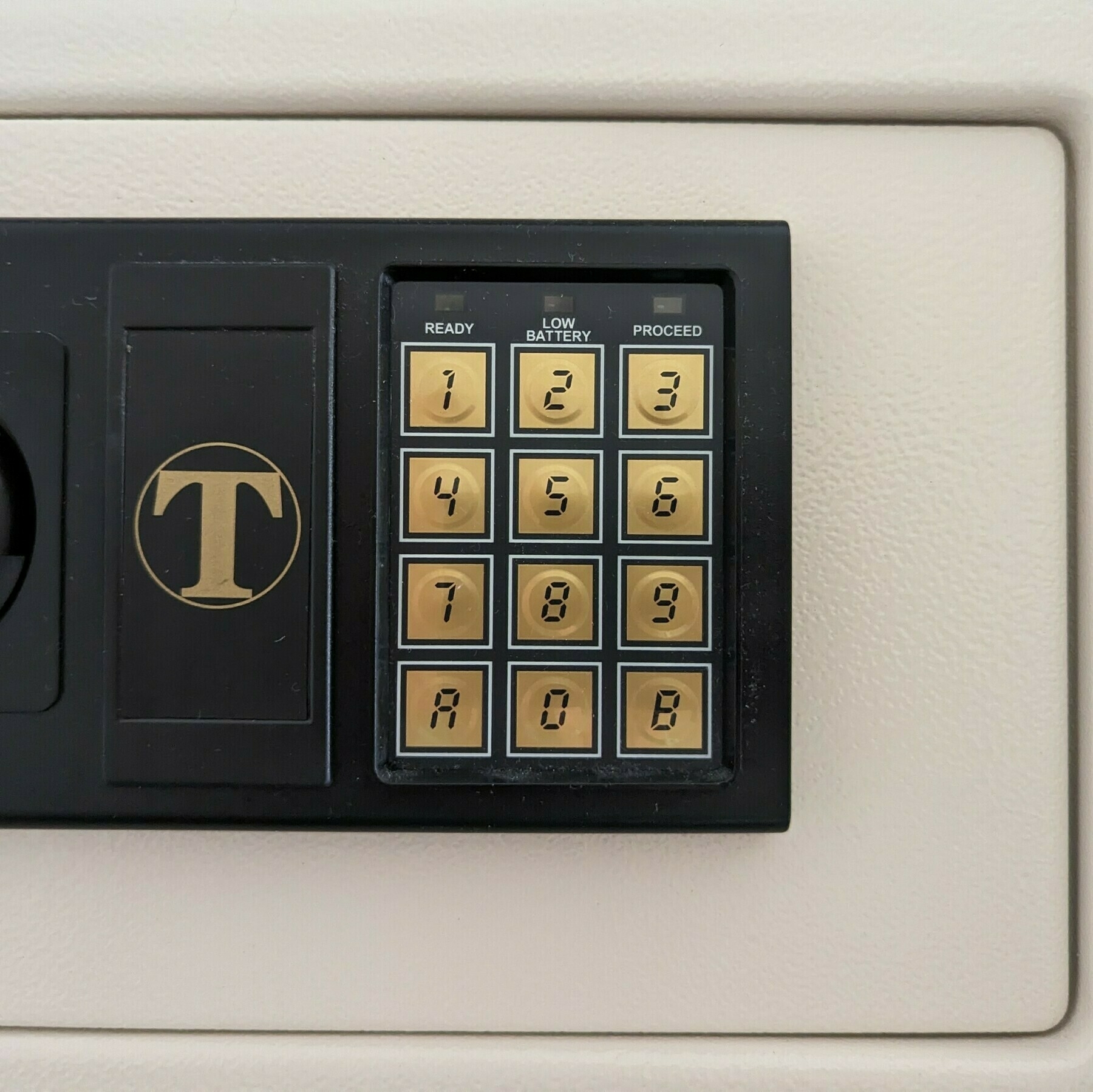 A numerical keypad on a safe with digits 0-9 and the letters A and B on either side of the zero in a seven segment display font. The left side of the B is a solid line, used to distinguish it from the loops of the B which are represented as they would appear on a seven segmented display. To the left is the safe's breading, which is a T in a circle.