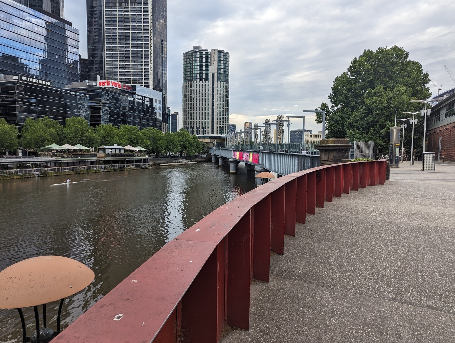 Concrete path with a metal railing beside a river leading to a bridge with skyscrapers in the background.