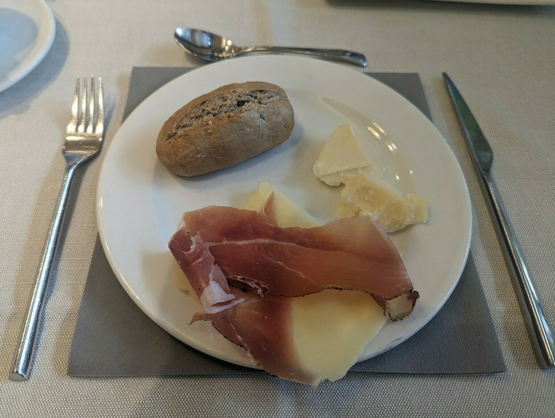 Plate on a place-mat with a wholemeal dinner roll, some parmesan cheese, slice of Jarlsberg cheese, and prosciutto. A silver knife and fork are beside it.