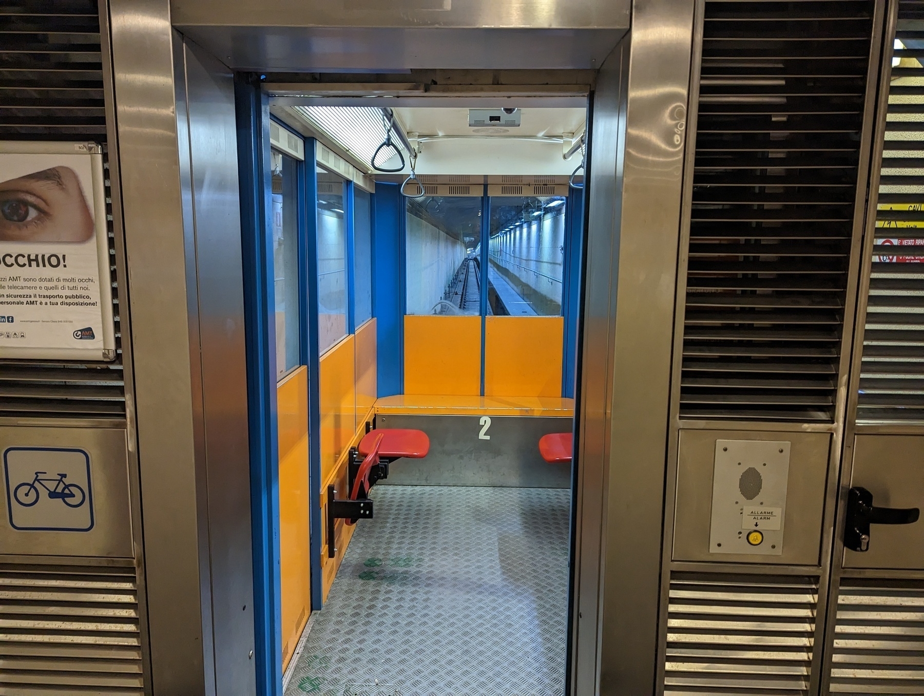 Empty lift car showing red fold-up seats and hand-loops on the ceiling. There are windows on all three sides of the inside of the car showing the corridor.