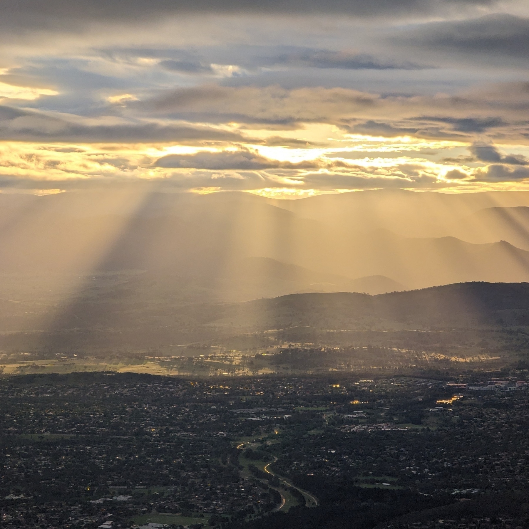 Aerial photo of a cloudy sunset over an urban area near the camera and hills in the distance, with sunlight streaming through the clouds and lighting the foothills.