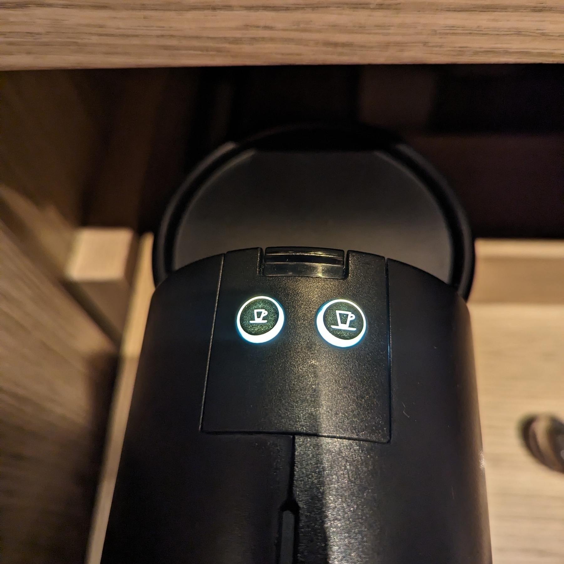 The top of a pod coffee machine, with two illuminated buttons, the power button that looks like a release for the water tank, and the top of the water tank itself