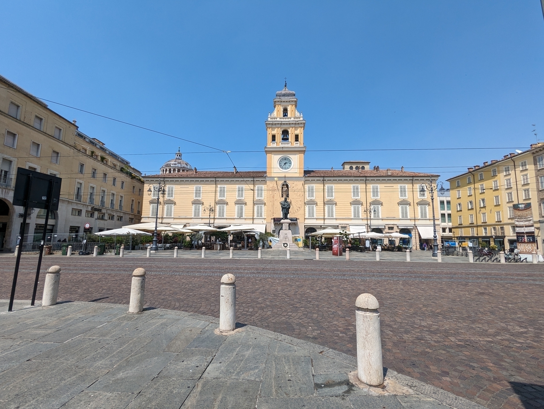 Front of the Palazzo del Governatore, a two story palace made of yellow stone, with a clock tower in the center. The front consists of umbrellas for outdoor dining of the cafe. In front of that is a street made of cobblestones, surrounded by bollards, and with trolly-bus overhead wires. The sky was sunny and cloudless.