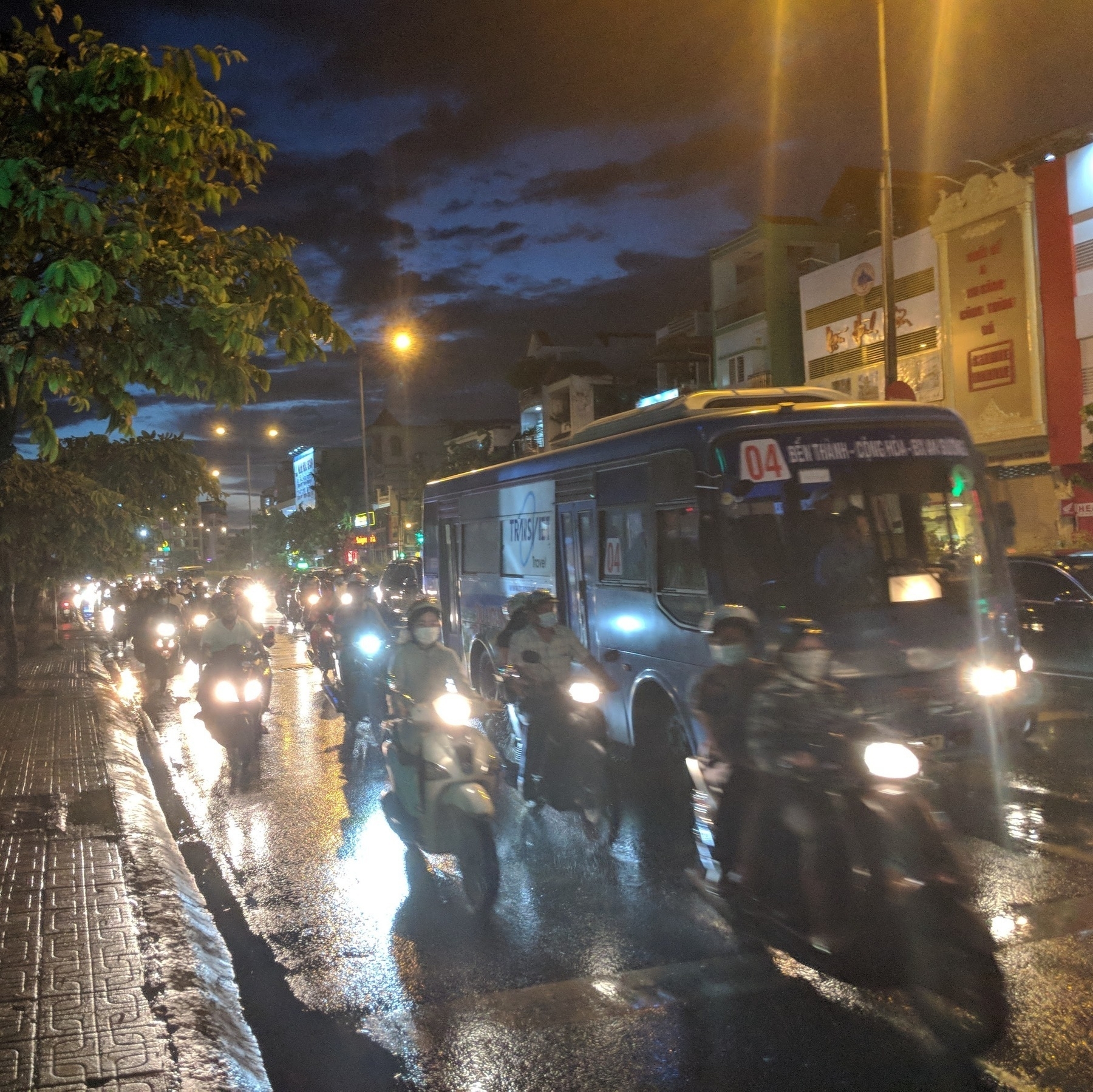 Motorcycles and scooter travelling along a road at night, with a bus travelling behind them. Head-lights and the streetlights are flaring.