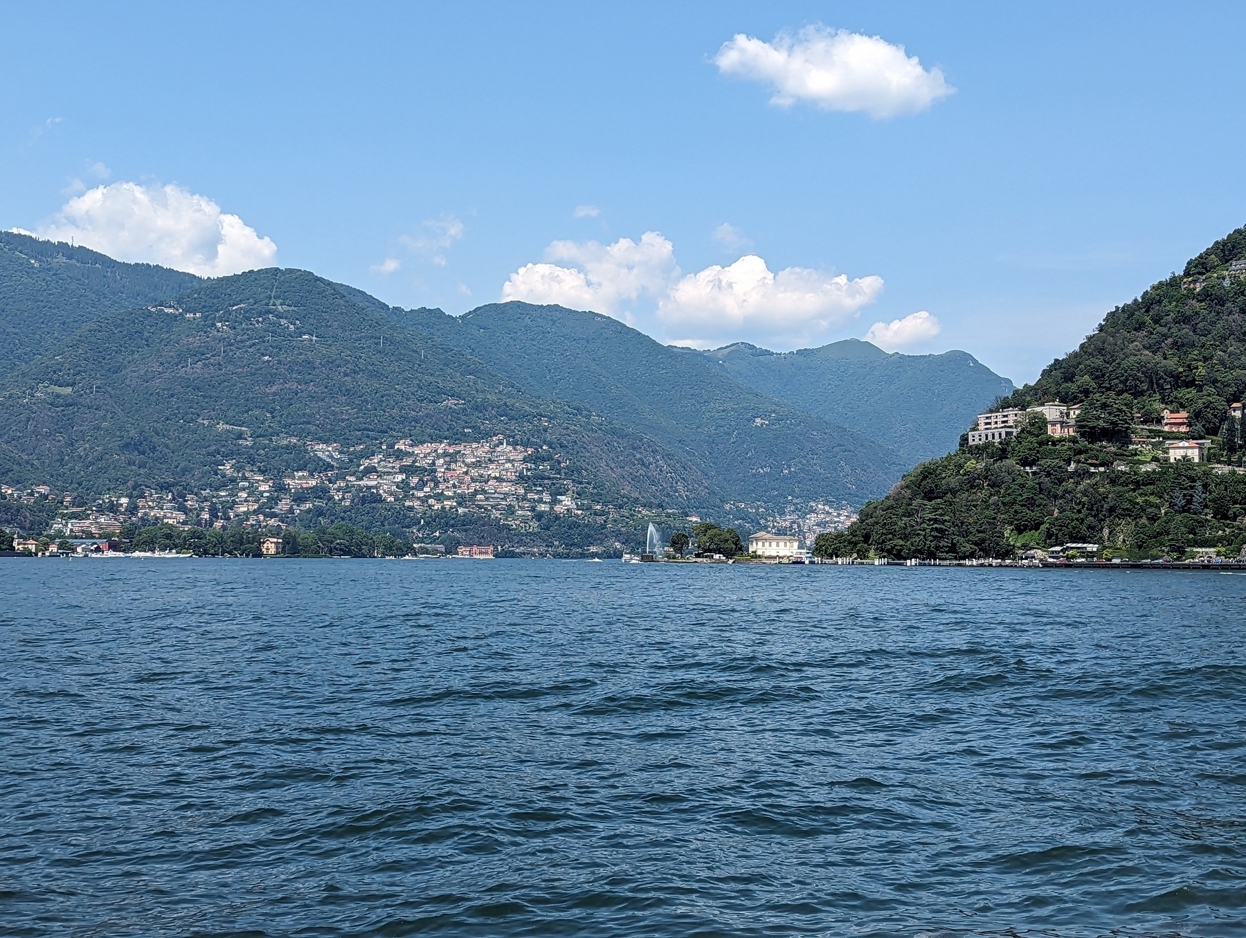 Lake Como, a large body of water with some small waves, with a fountain in the middle distance, with green mountains in the background, with a slightly cloudy sky