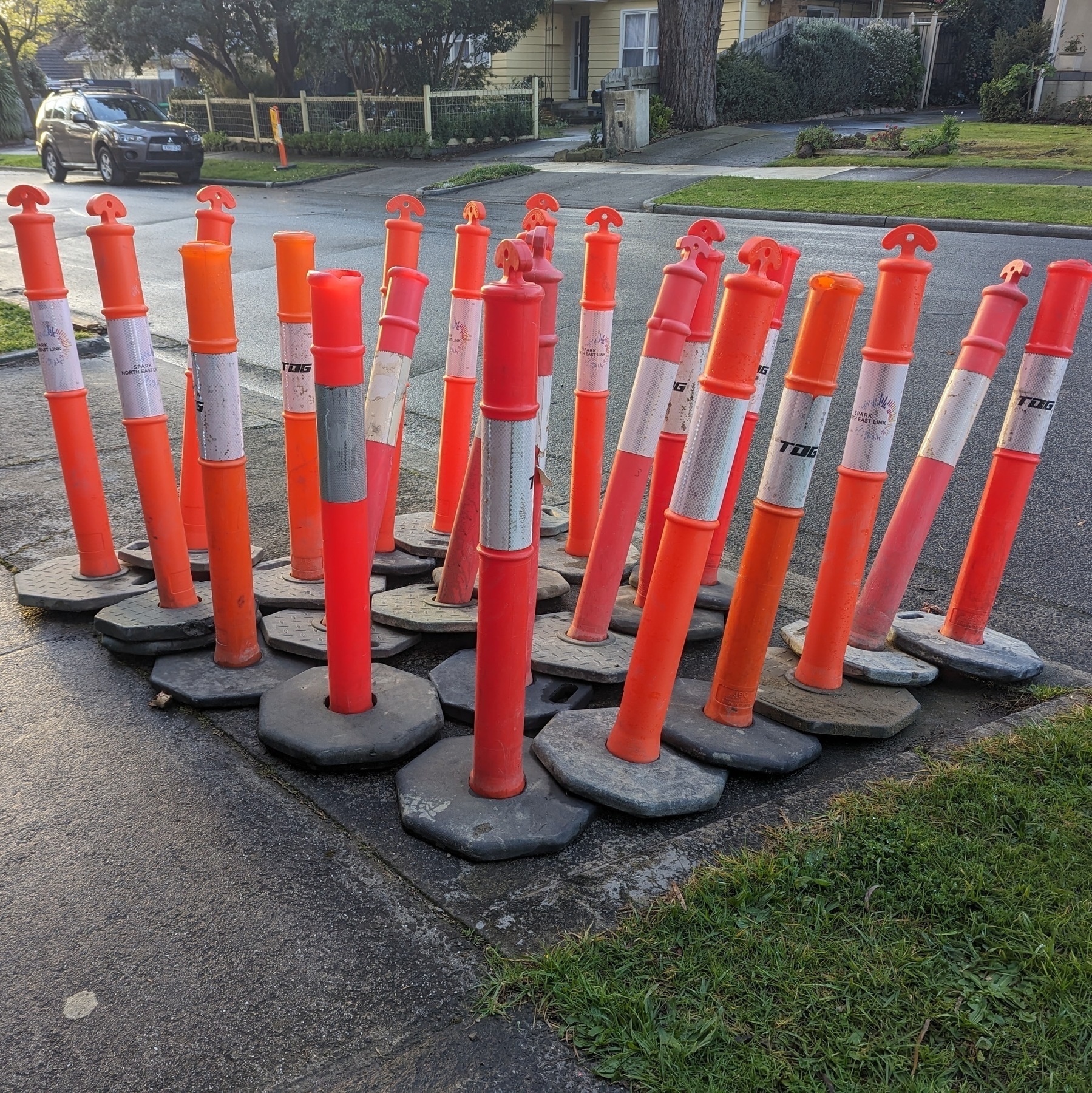 A group of orange traffic cylinders, sort of like cones but about waste high, on the side of a road with a black car in the background