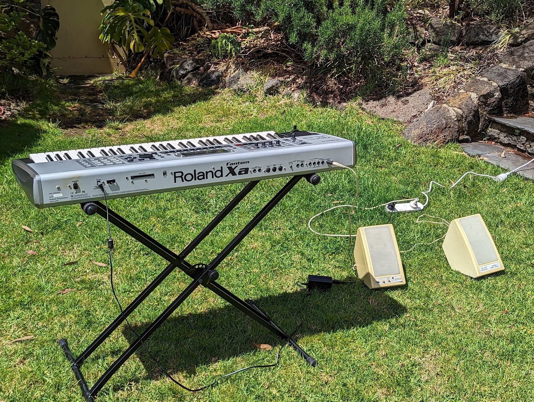 A keyboard synthesiser on a keyboard stand in a garden, connected to two small, yellowed PC speakers which are sitting on the ground to the right