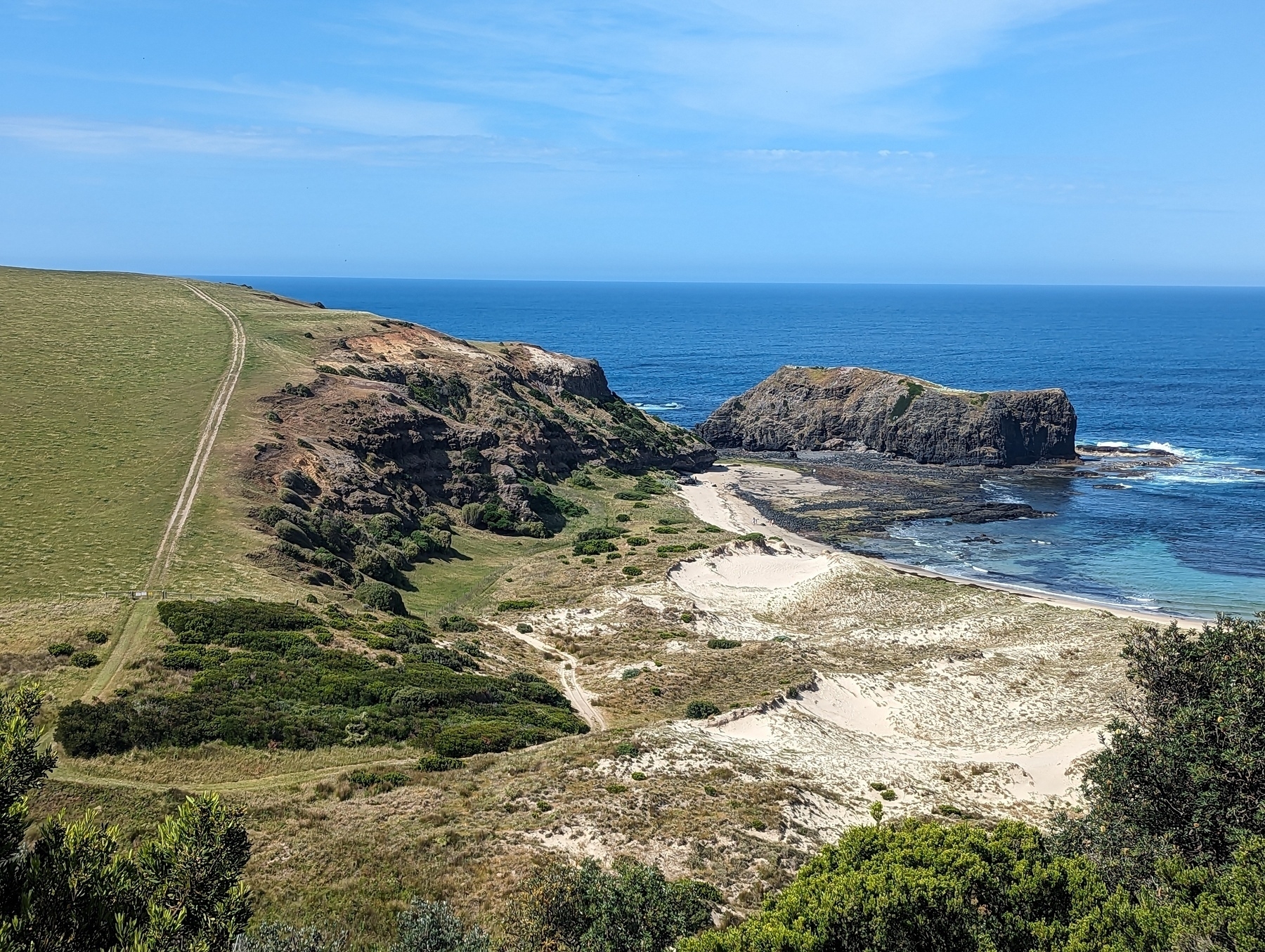 An elevated photo of a beach with the sea on the right, a cliff in front, a road going along the ridge, and a green field on the left