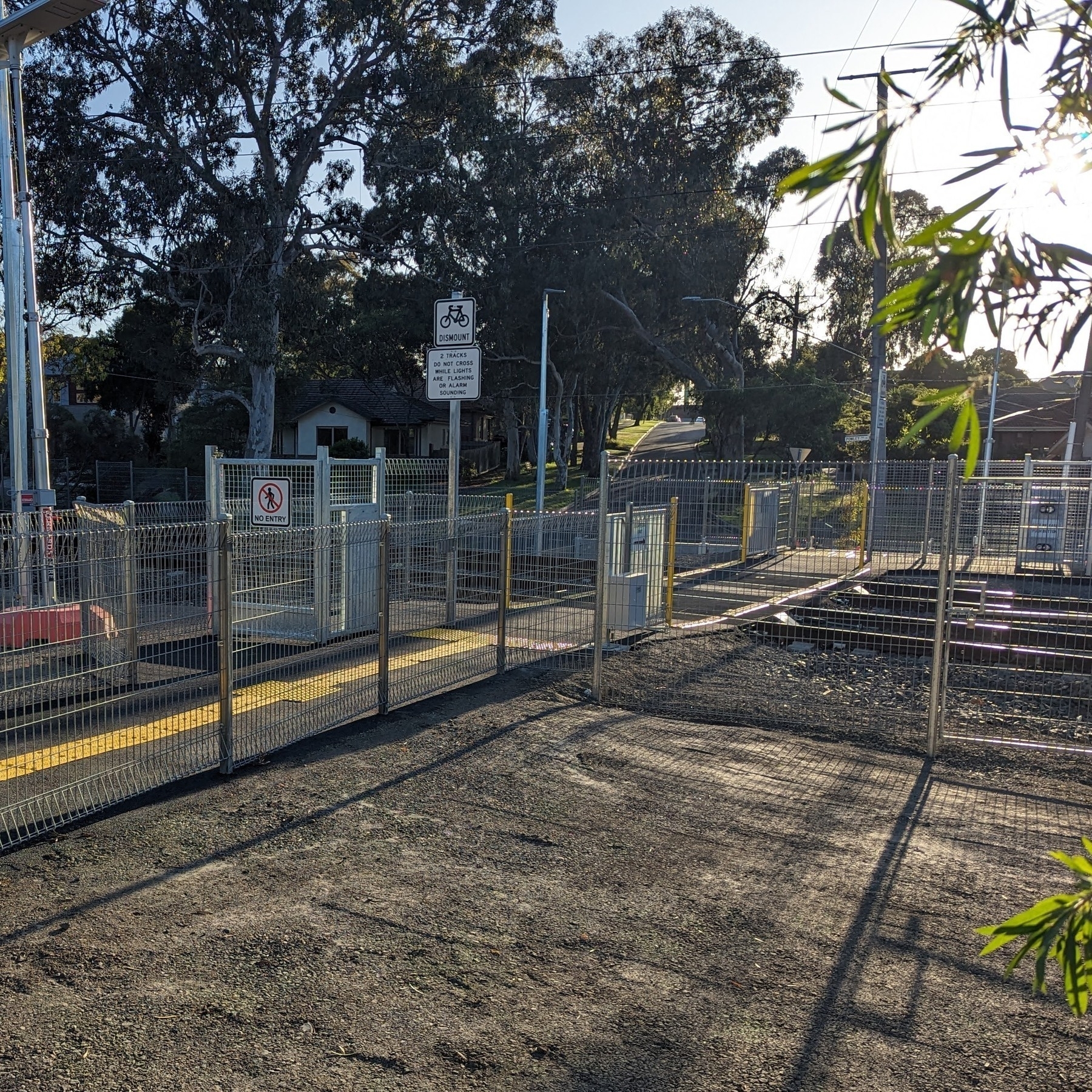 A pedesterian rail crossing with feces and automated gates that will close when a train passes by. This is backed by suburban streets and trees.