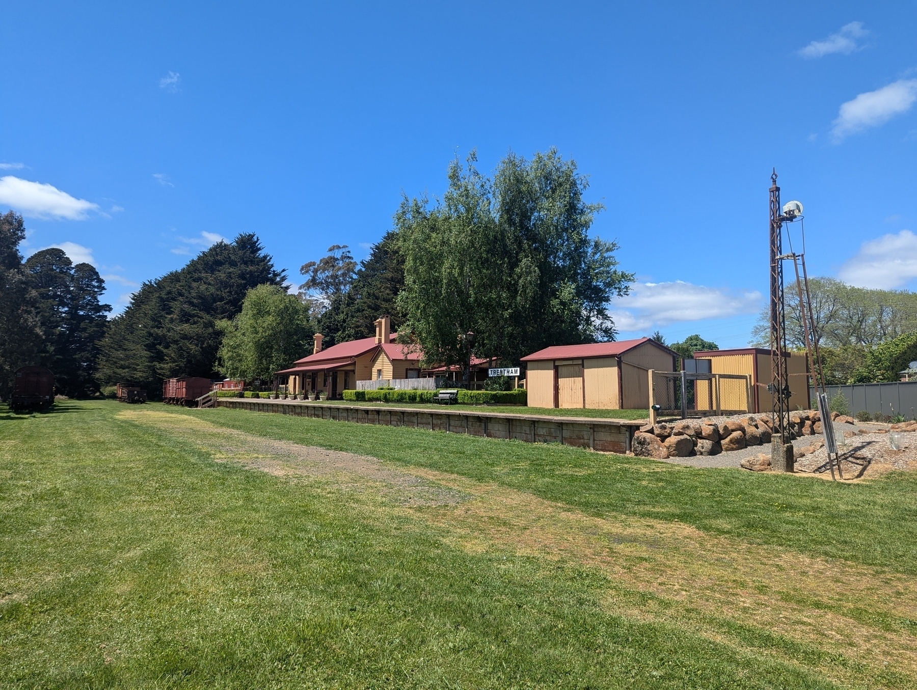 A former rural train station surrounded by lush greenery and clear blue skies, with old rolling stock and an old semaphore signal post.