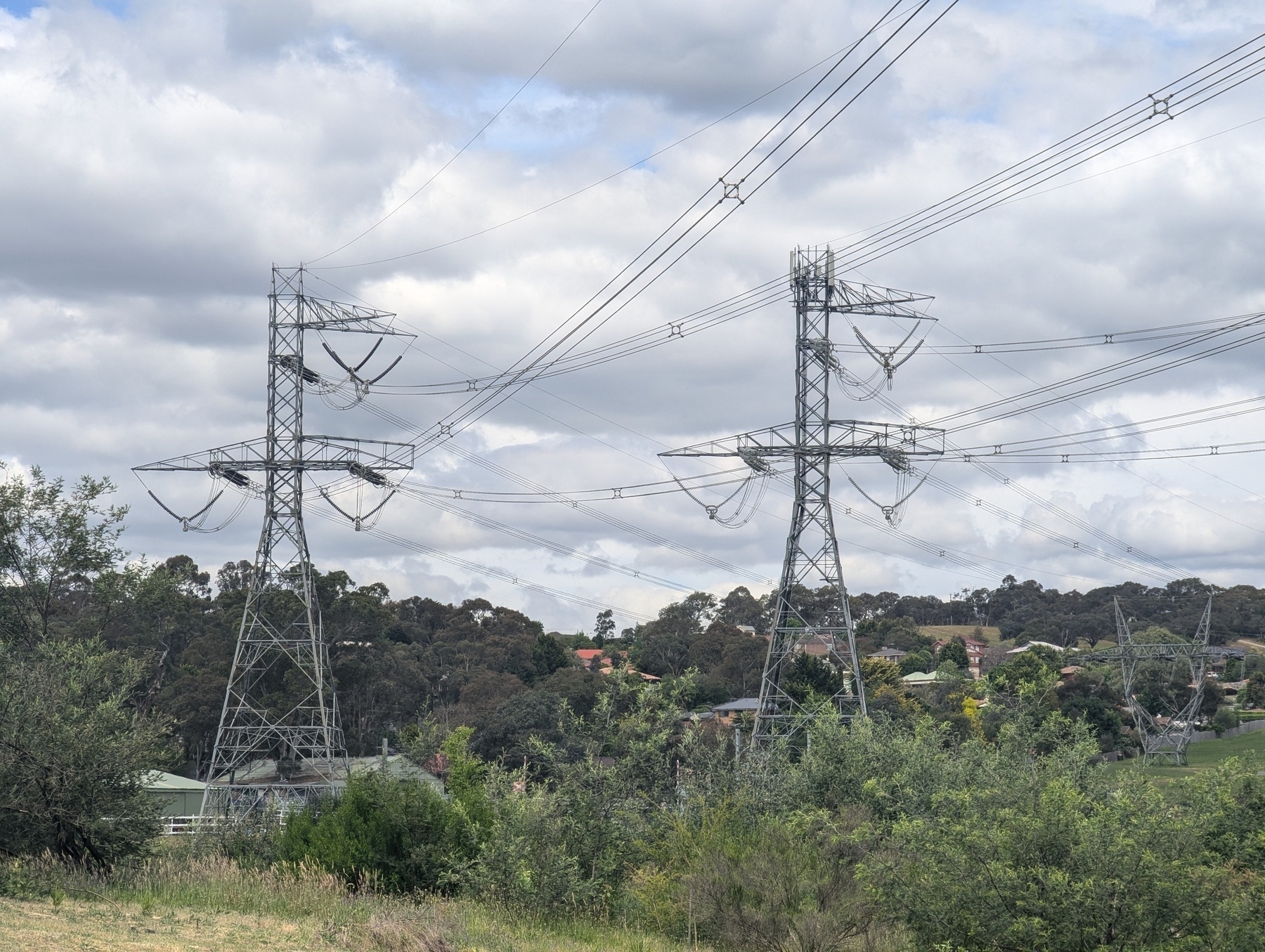 Two large metal electricity pylons stand in a grassy area with trees in the background under a cloudy sky.