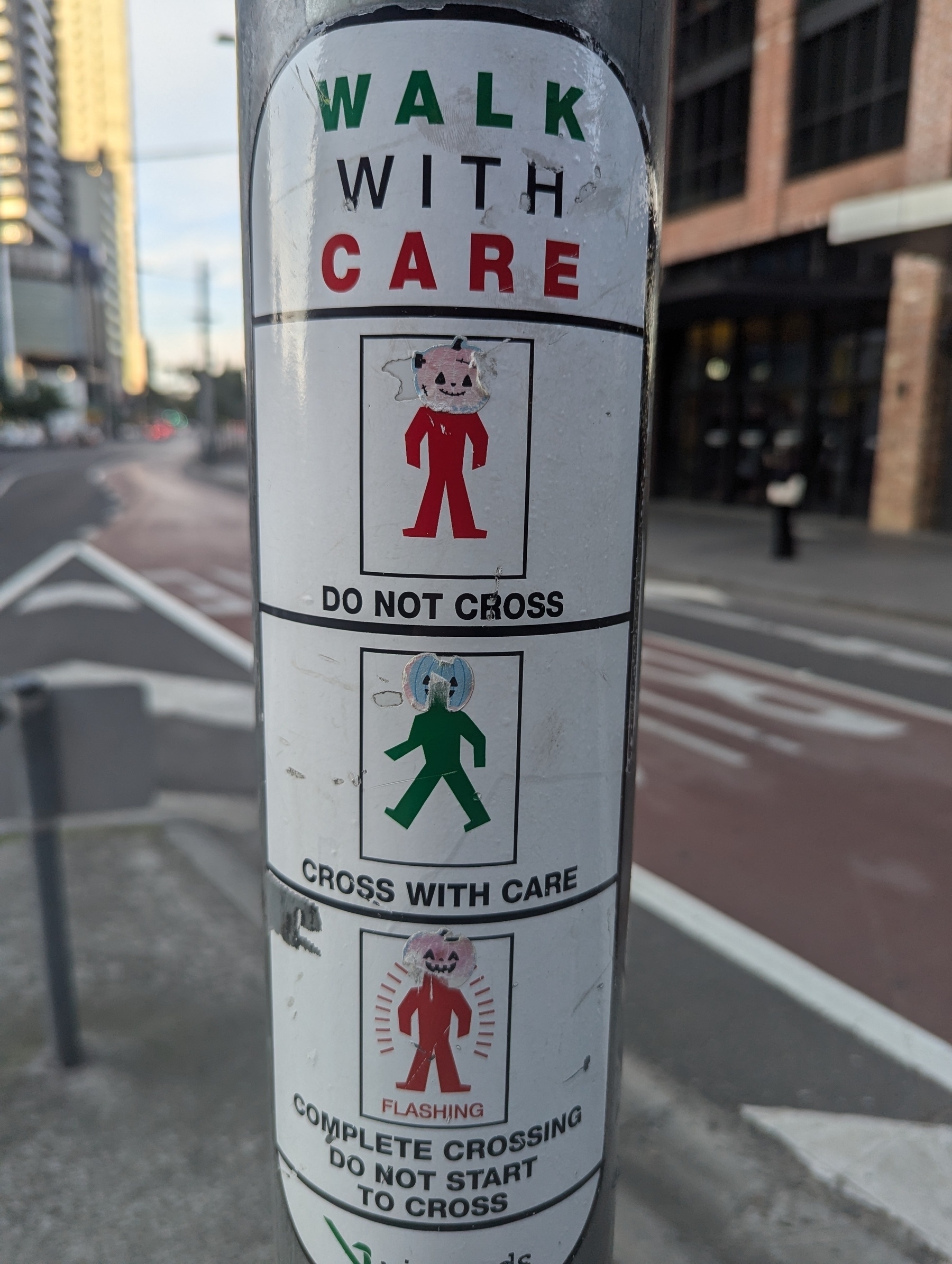 A street pole features a sign with pedestrian crossing instructions, using humorous cartoon characters with jack-o'-lantern placed over their faces to indicate "Do Not Cross," "Cross With Care," and "Complete Crossing Do Not Start to Cross."