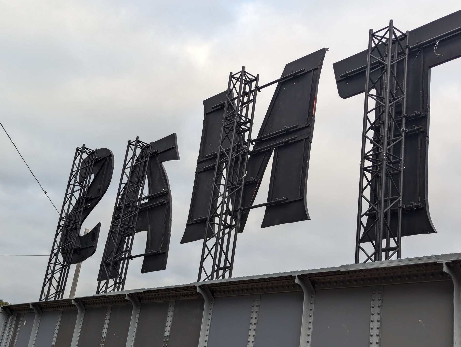 Large letters TMAS mounted on a bridge structure appear reversed against a cloudy sky backdrop.