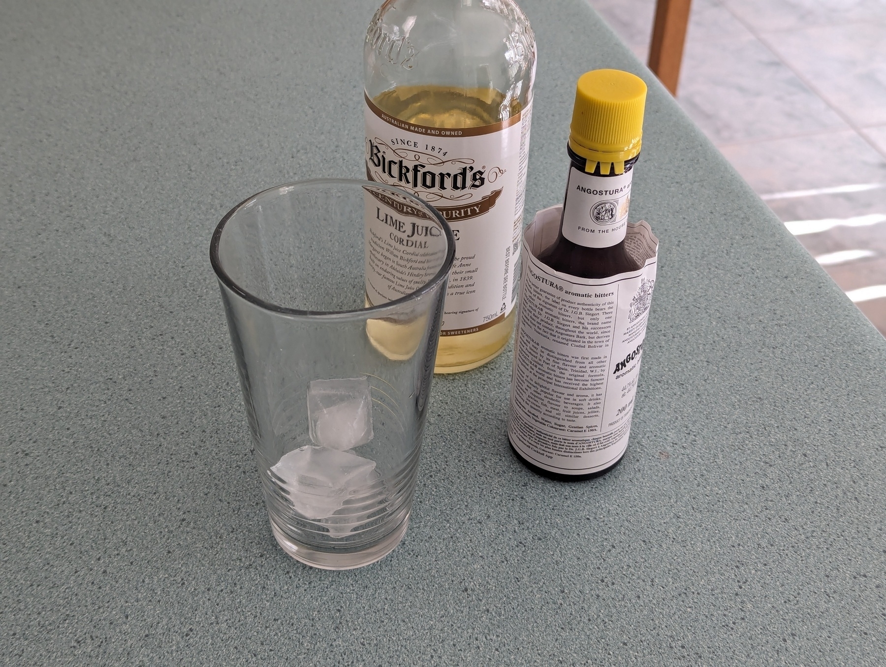 An empty glass on a bench containing ice cubes, with bottles of lemon and lime cordial and bitters behind it.
