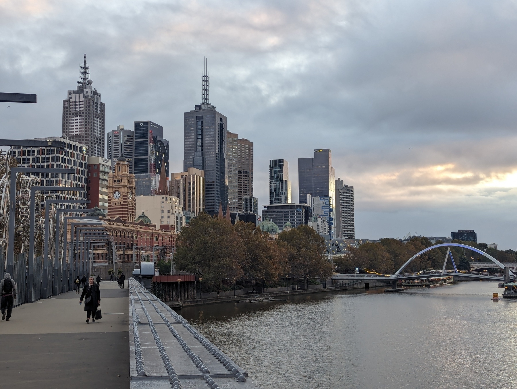 Photo of the Melbourne skyline, facing Flinders St. station and the CBD, with the pedestrian bridge on the right, as taken from a bridge with an iron railing over the Yarra River.