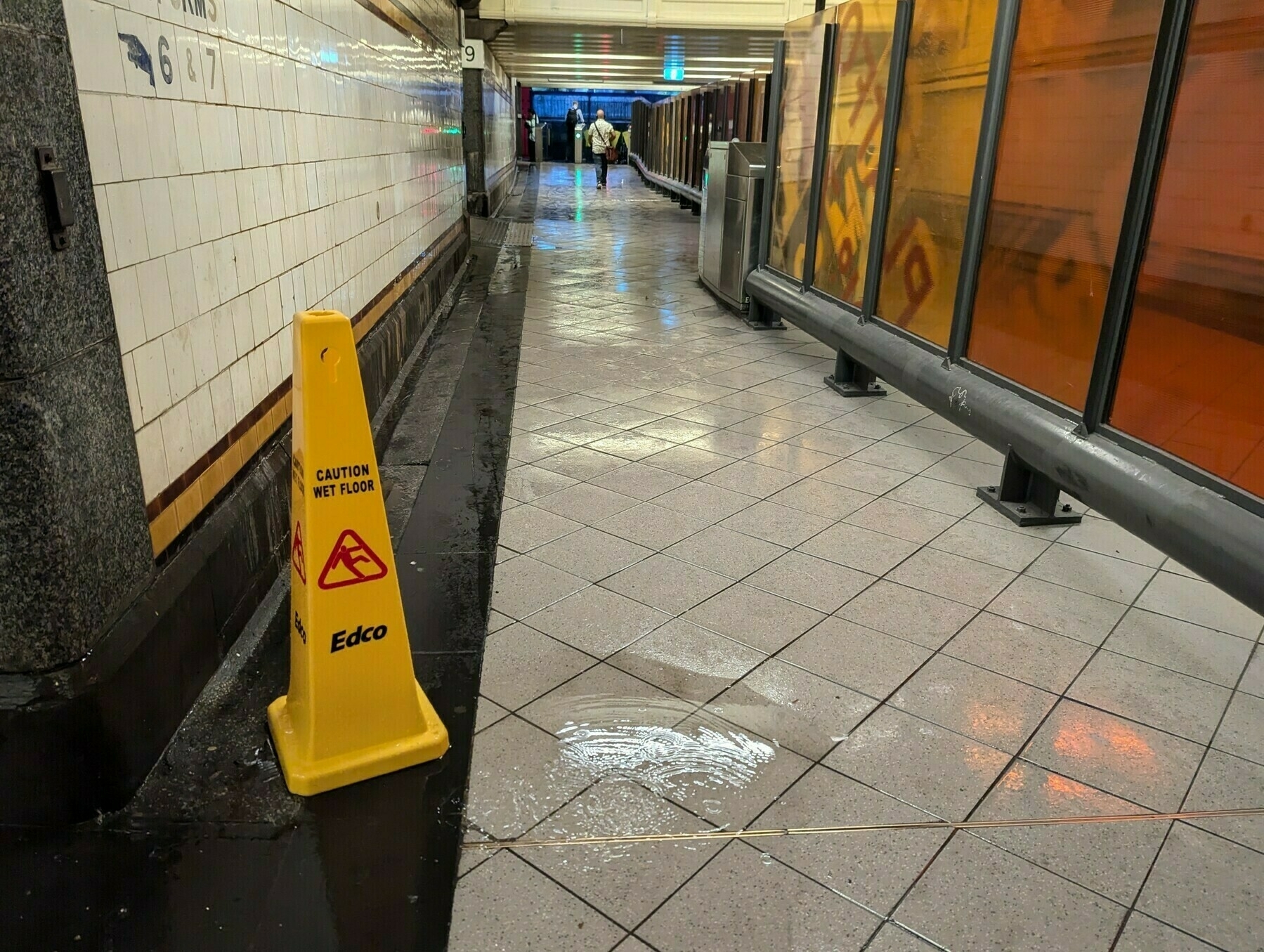 Auto-generated description: A wet floor caution sign stands in a tiled underground walkway with puddles on the floor.