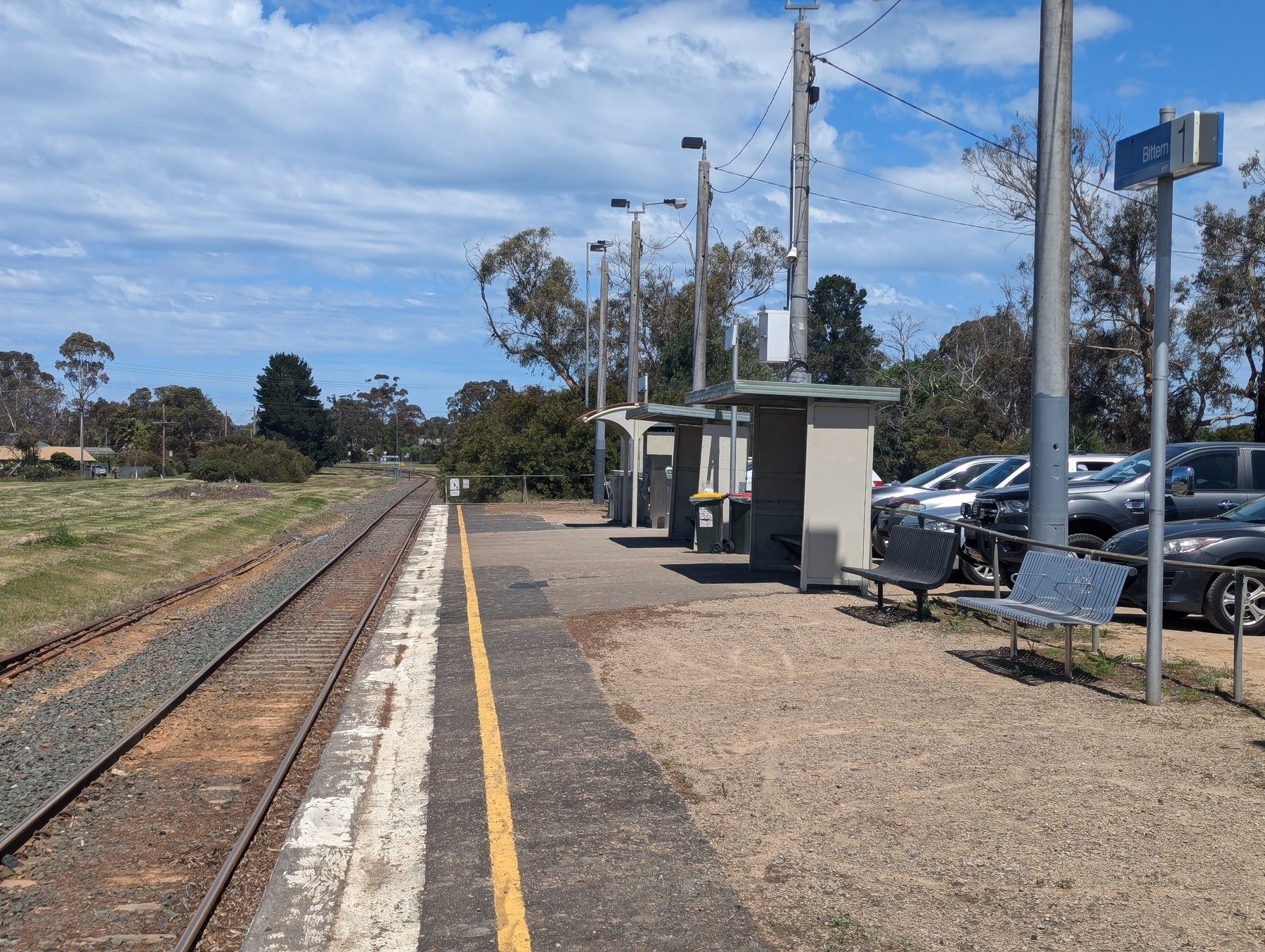 A small train station platform is situated alongside a single railway track, with nearby parked cars and a partly cloudy sky overhead.