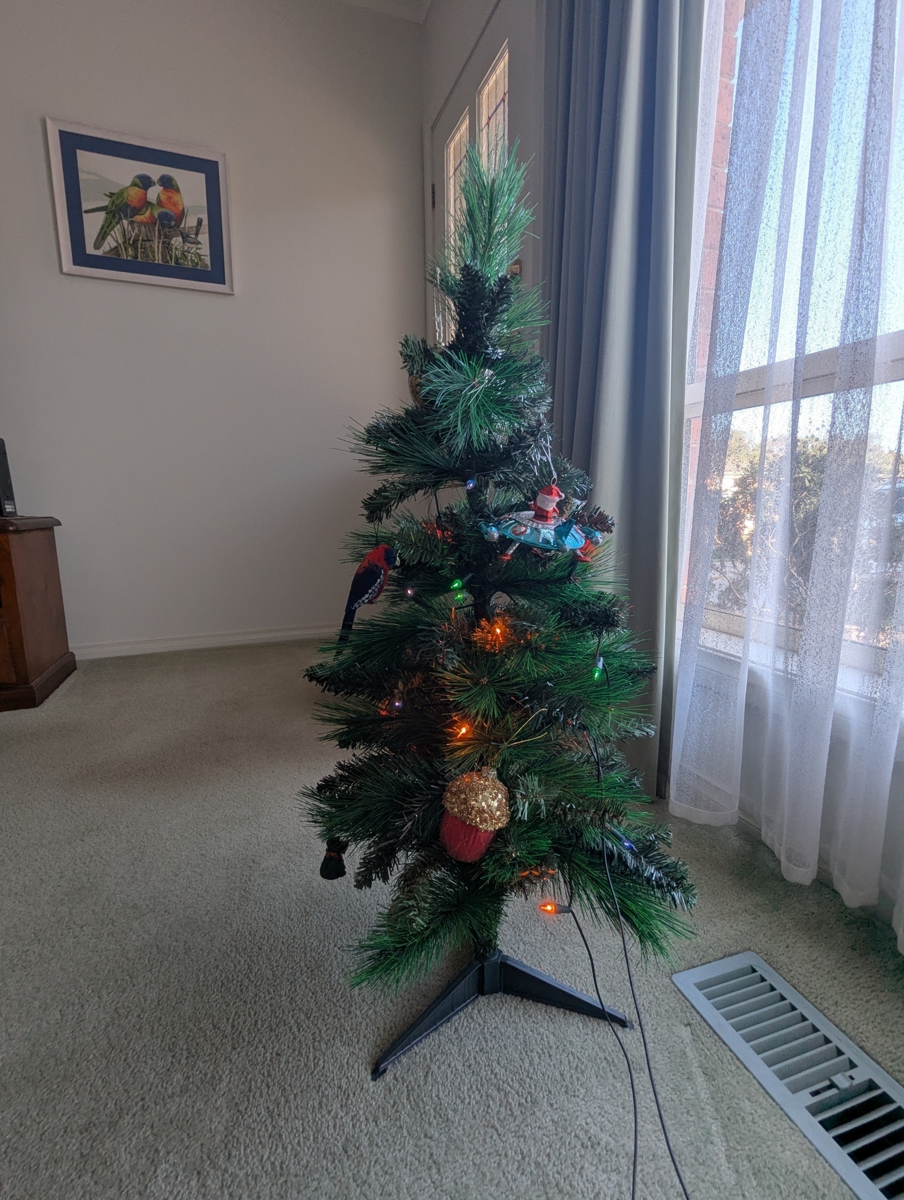 A small decorated Christmas tree stands in a living room near a window with sheer curtains.