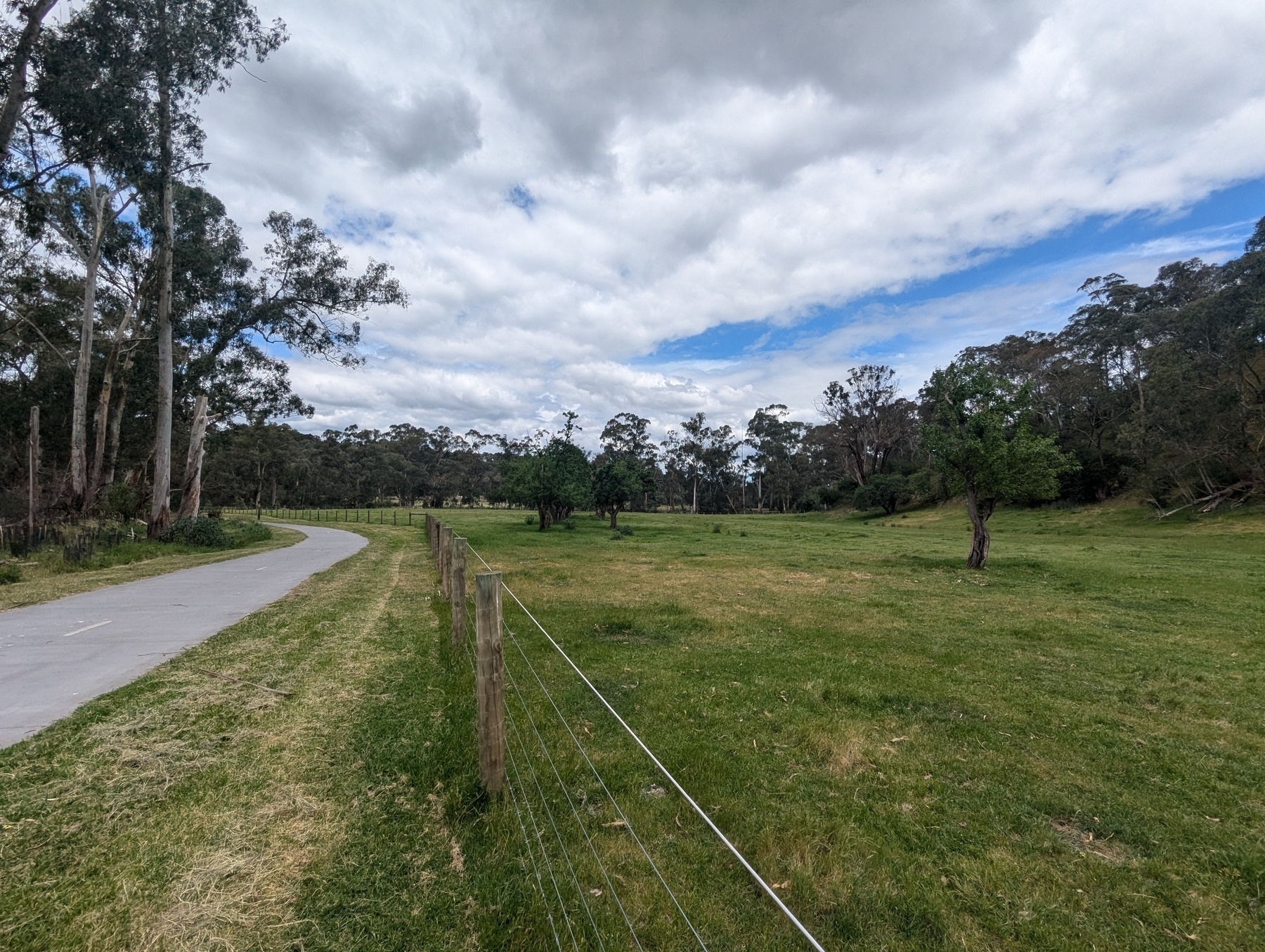 A paved path runs alongside a fenced grassy field with trees under a cloudy sky.