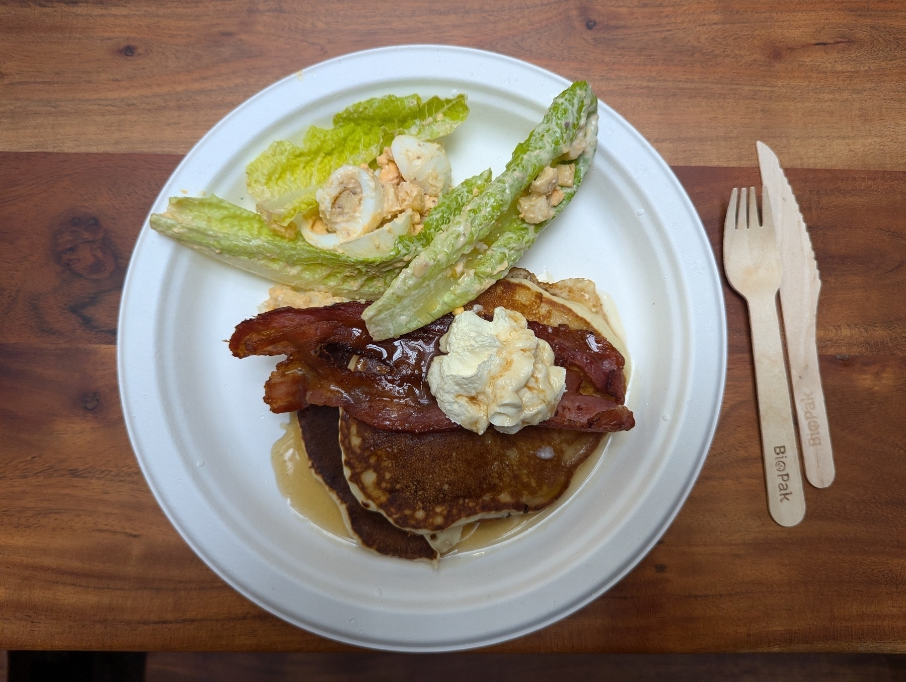 A plate with pancakes, bacon, whipped cream, drissled with maple syrup, with a side of Ceaser salad and egg. Wooden knife and fork on the right.