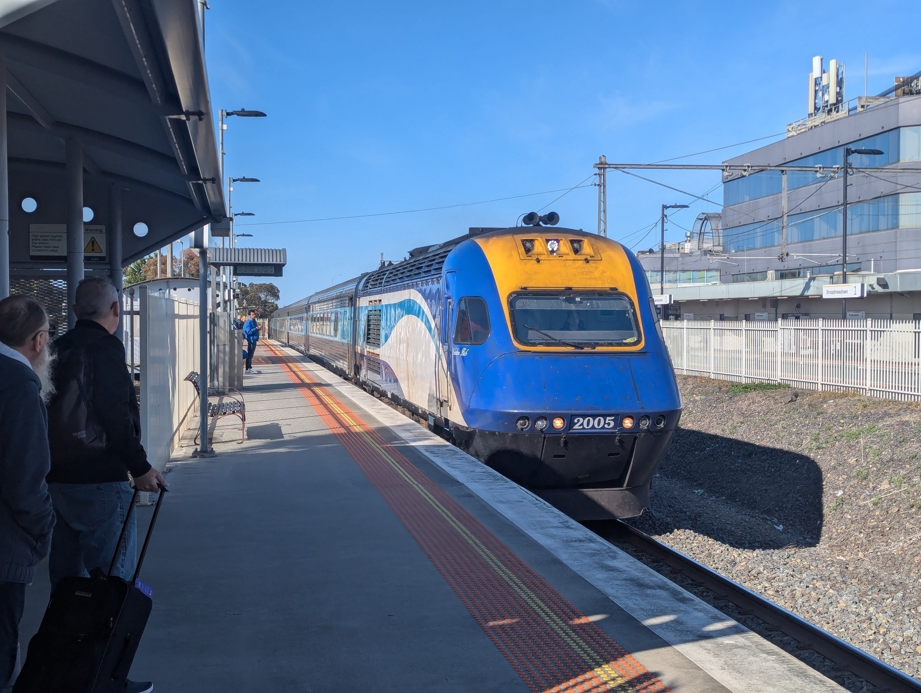 Autogenerated description: A train with a blue and yellow front is arriving at a station platform under a clear blue sky, with people waiting nearby.