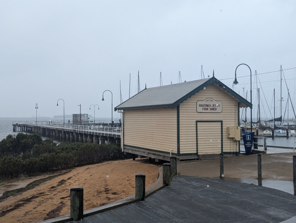 A small building labeled Fire Shed is situated near a dock with numerous sailboats in the background on a cloudy day.