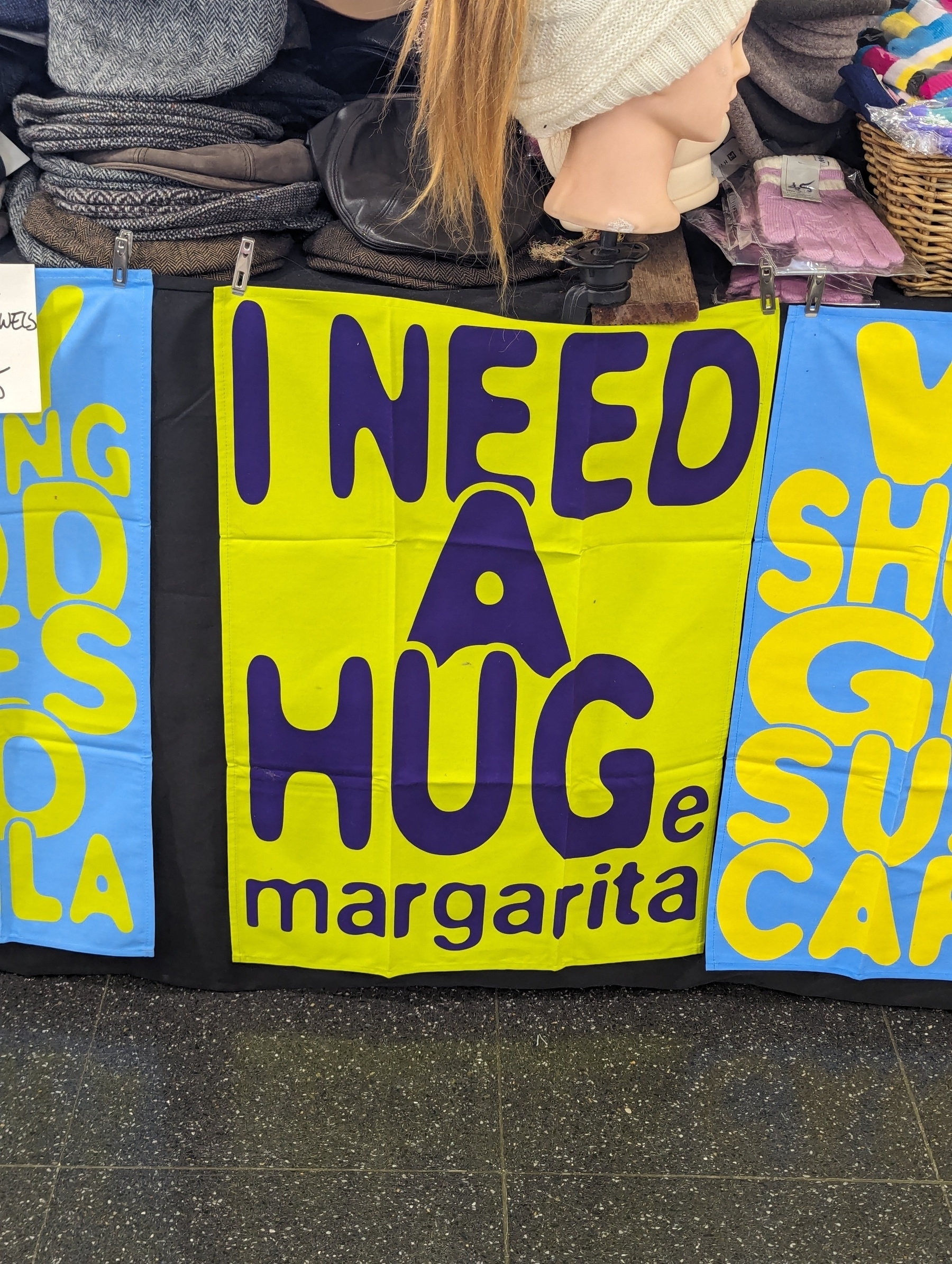Yellow tea-towel on a display with the phrase 'I need a hug' in large letters, followed by 'huge margarita' in small letters. The first three letters of 'huge' were borrowed from the word 'hug'.