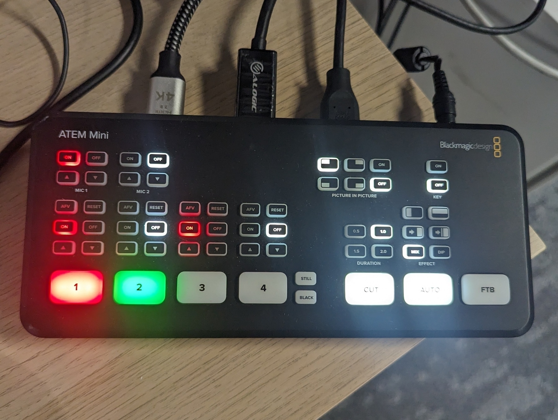 A Blackmagic Design ATEM Mini on a wooden desk with some of the buttons on the front panel illuminated red, green and white. Cables are plugged into the back.