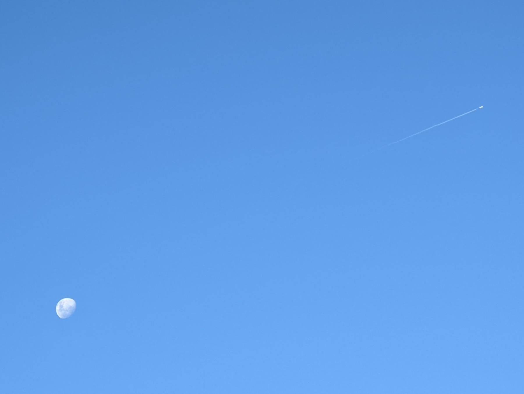 A clear blue sky, with the moon at the lower-left side of the frame, and a plane with a vapour trail near the right