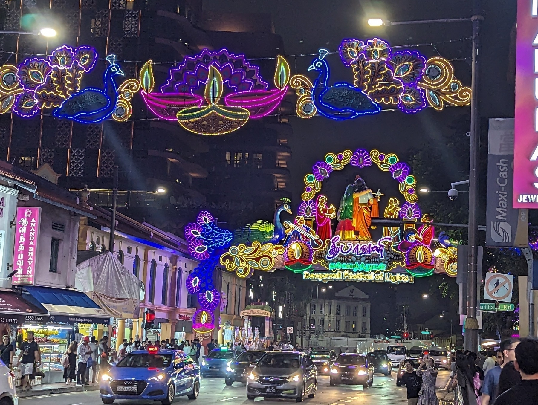 A city road at night, with cars driving towards the camera, and yellow, purple, and blue lights strung up above the street, featuring peacocks and Rangoli decorations celebrating Diwali