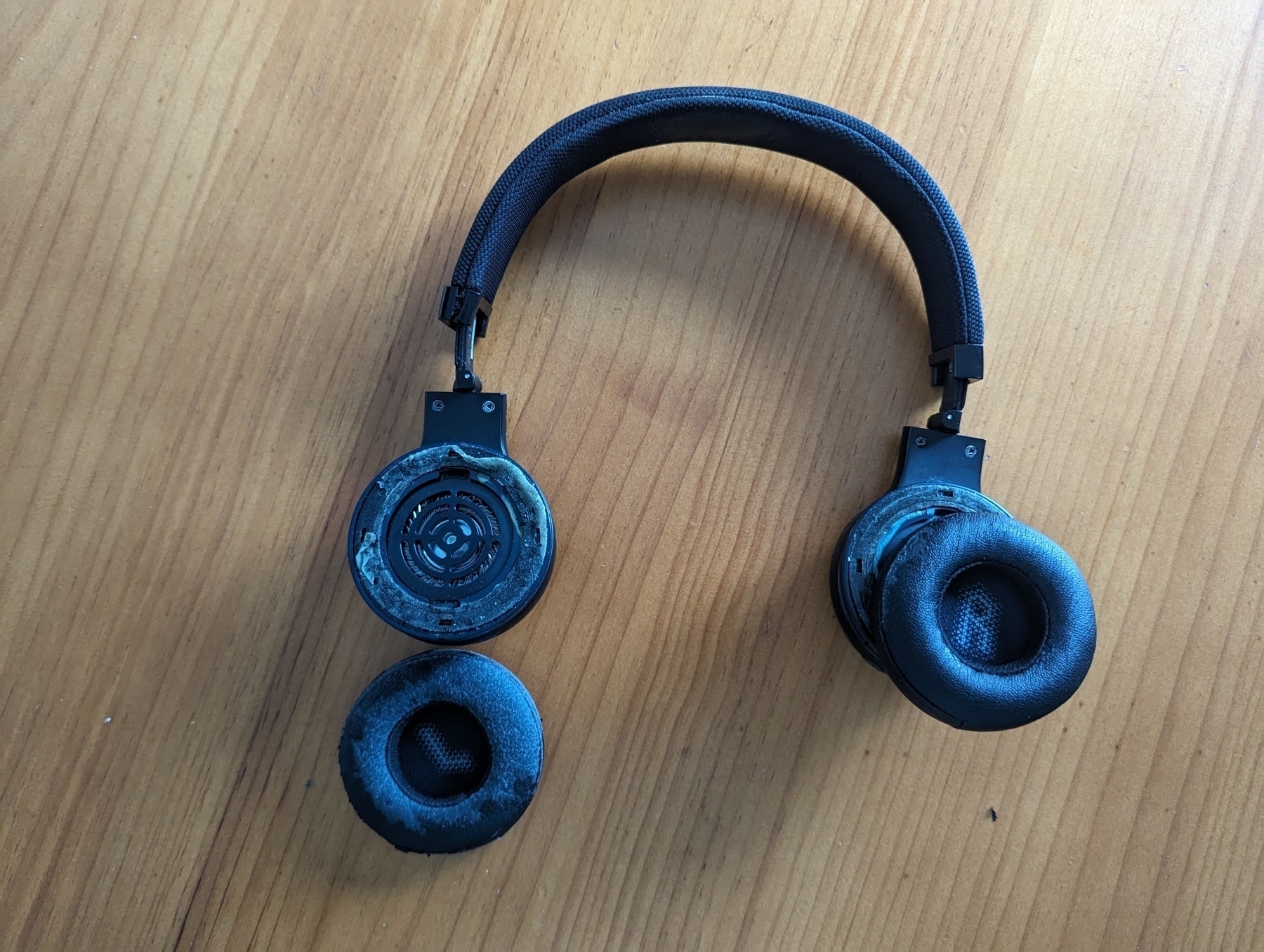 A black bluetooth headphone on a table, with the left cup fallen off exposing the speaker, and the right cup slightly removed from it's original position