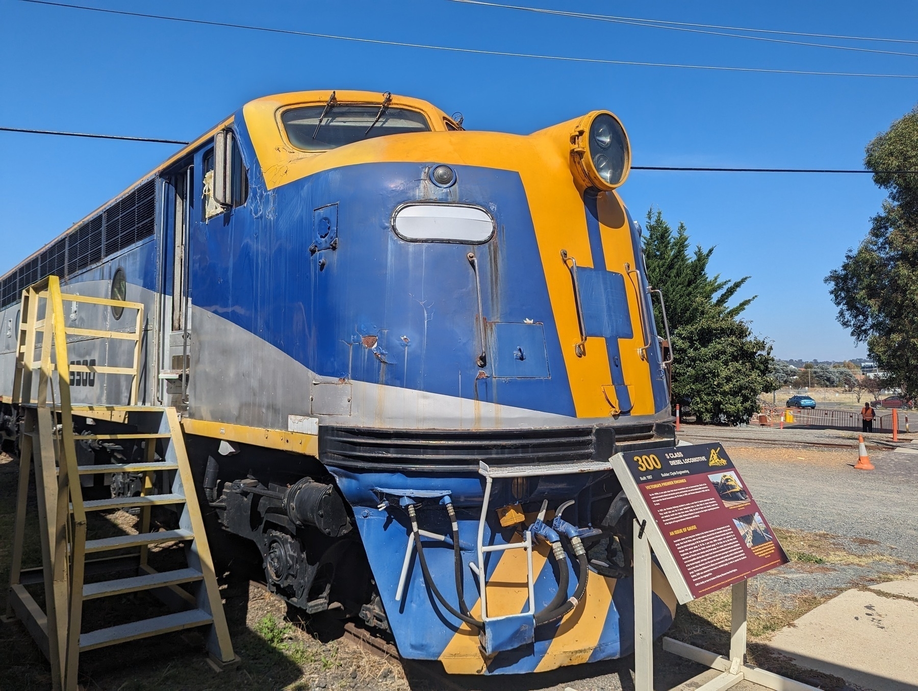 A vibrant blue and yellow vintage train engine is displayed outdoors with informational signage and a staircase leading up to it.