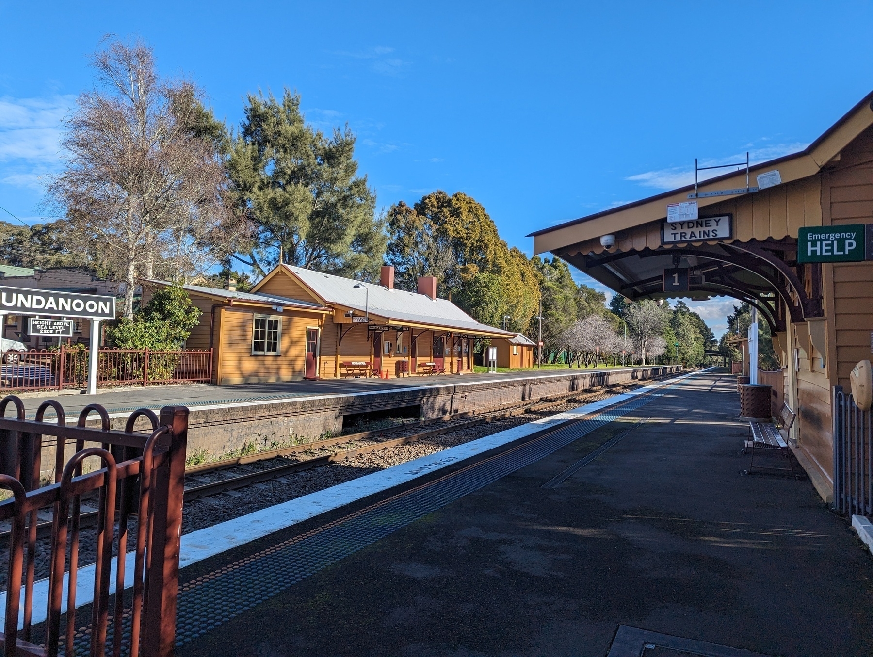 Auto-generated description: A quaint train station platform with a sign that reads Bundanoon is shown, surrounded by trees and blue skies.