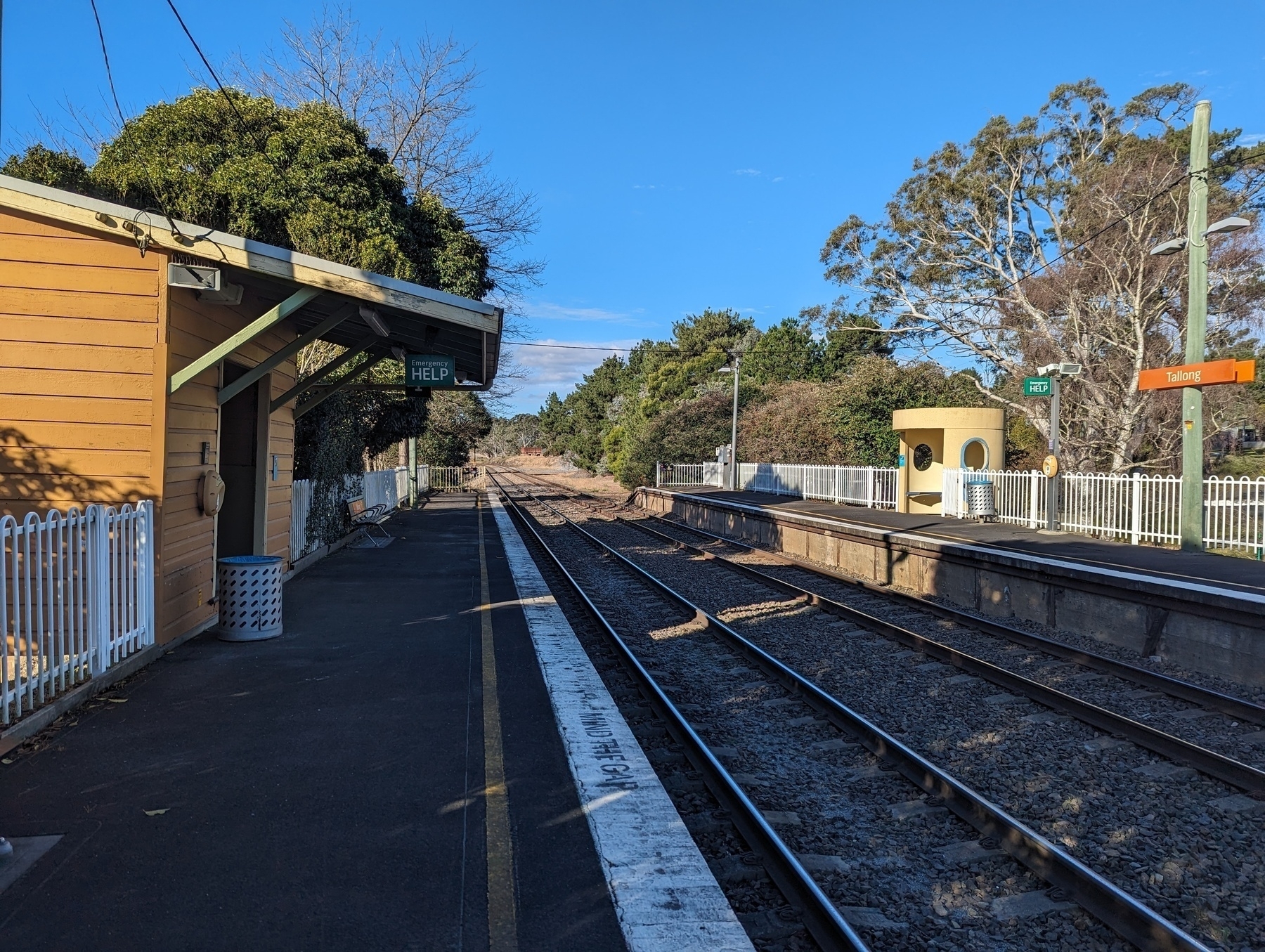 Auto-generated description: A quiet train station platform is shown with tracks stretching into the distance and surrounded by trees.