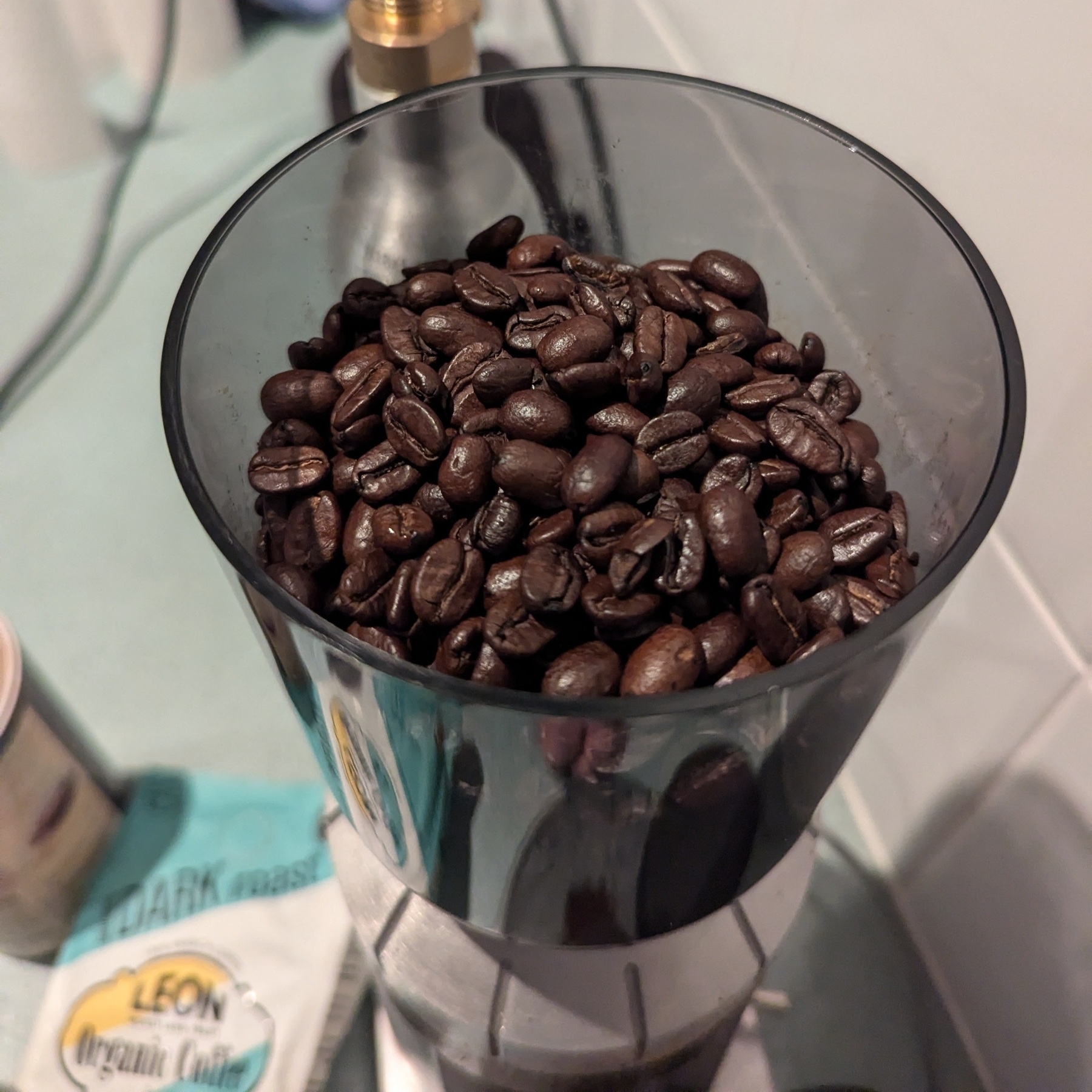 Auto-generated description: The hopper of a coffee grinder filled with roasted coffee beans, surrounded by various items on a bench including an empty bag of Leon coffee and what appears to be a small container of drinking chocolate.