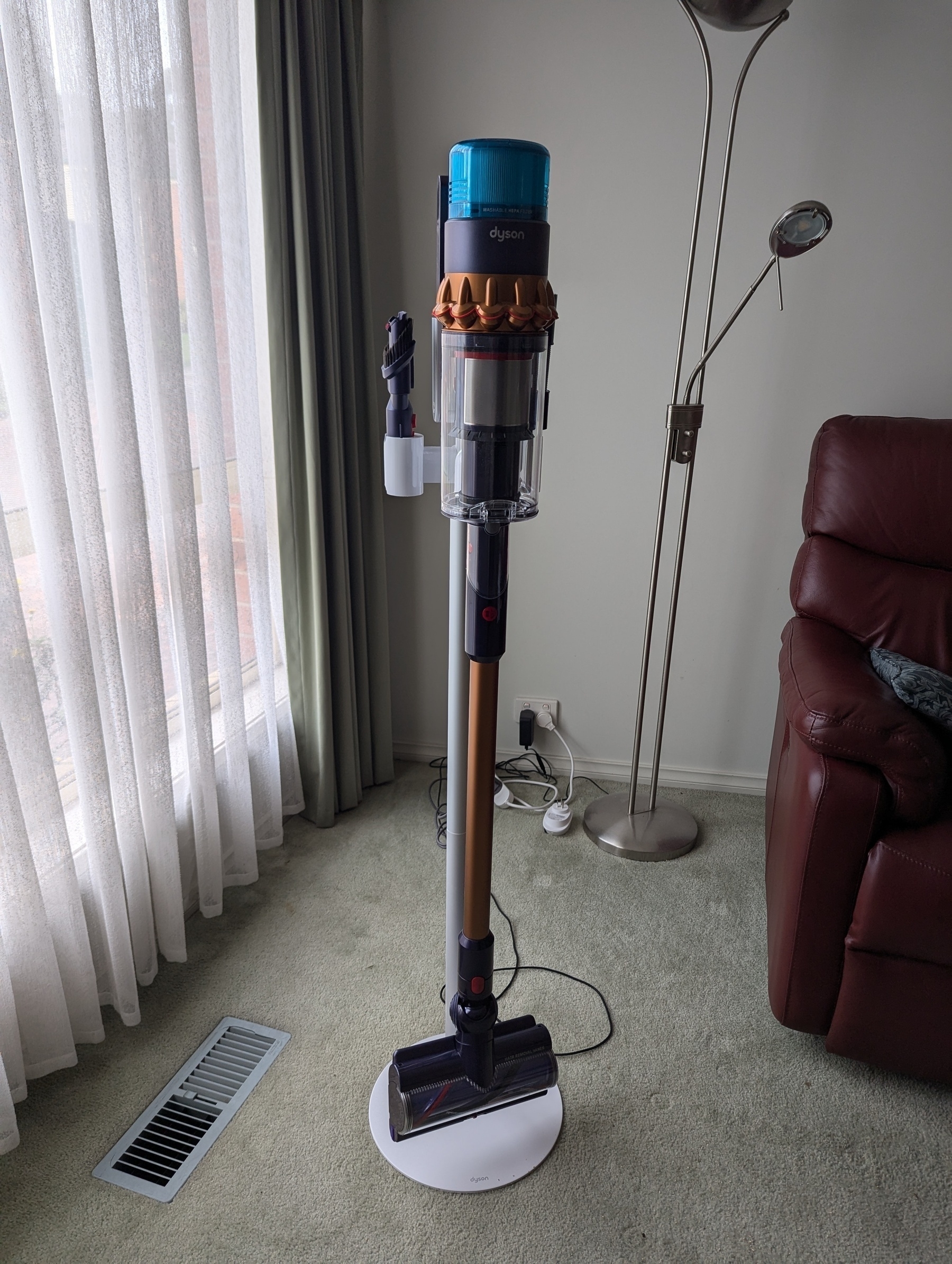 A cordless vacuum cleaner standing upright on a charging dock in a living room with a floor lamp, curtain, and a couch in the background.