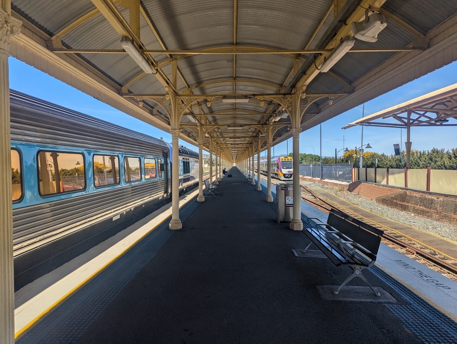 Auto-generated description: A train is parked at a long, empty station platform with a covered walkway and bench.