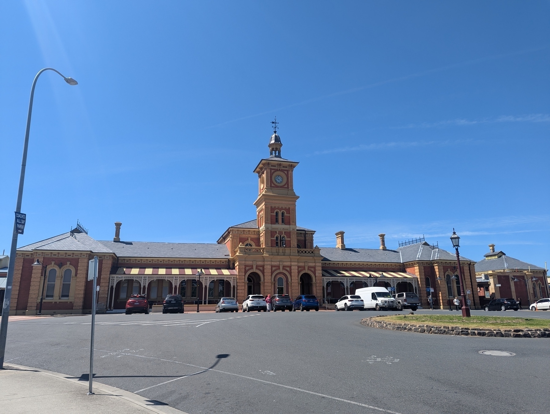 Auto-generated description: A historic railway station with a central clock tower is set against a clear blue sky, surrounded by cars and a lamp post.