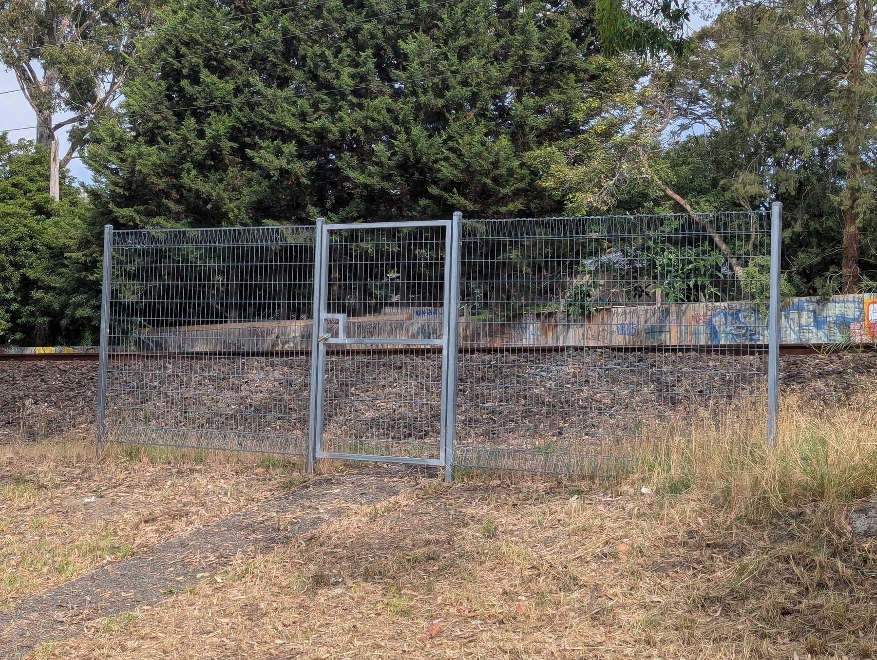 A wire fence, with a locked gate yet gaps opened on the side, stands in front of a railway track, surrounded by trees and grass.